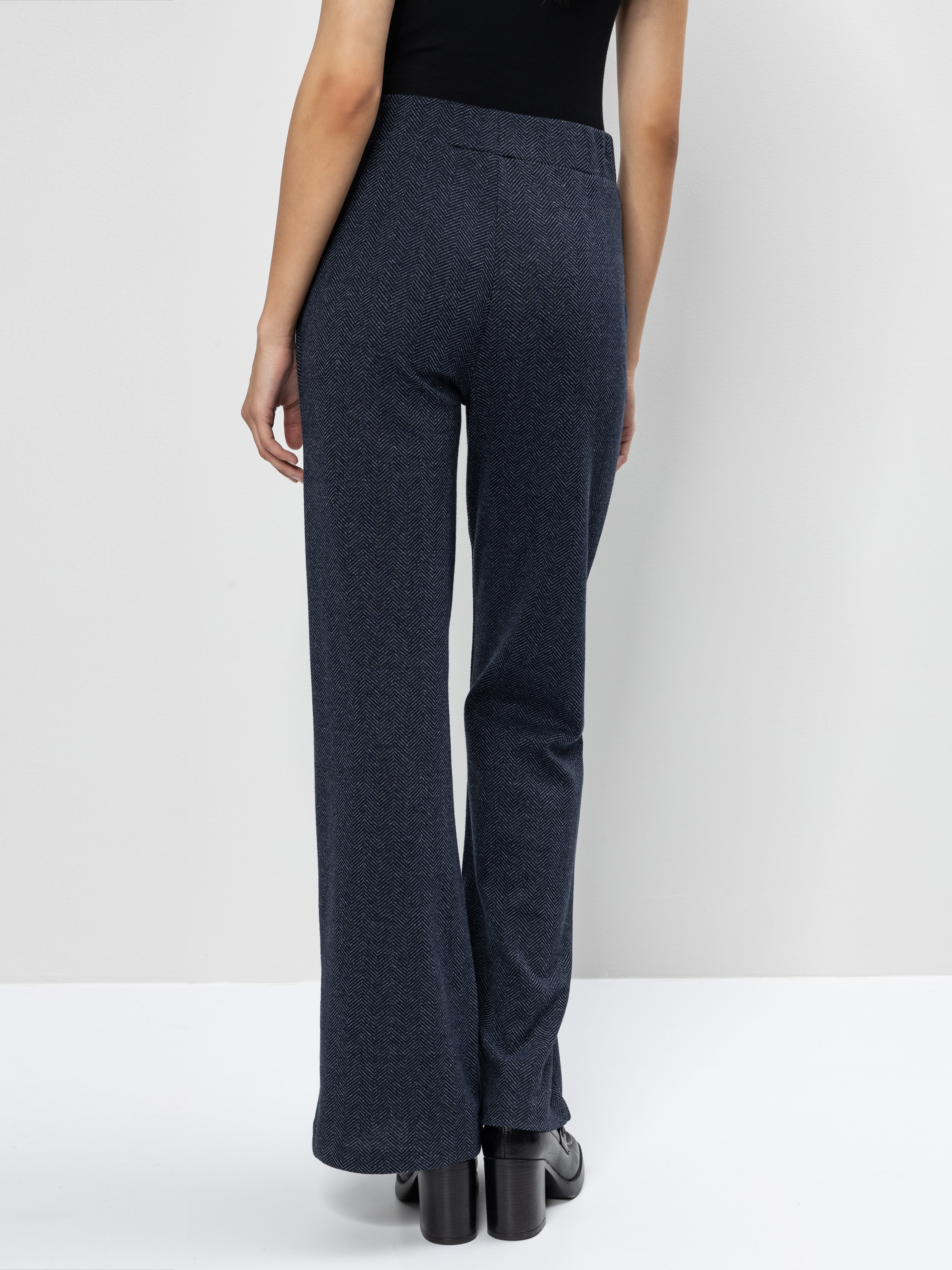 Eine Frau, die eine dunkelblaue, weit geschnittene Hose mit hoher Taille und schwarze Plateauschuhe trägt, steht mit dem Rücken zur Kamera vor einem schlichten hellen Hintergrund.