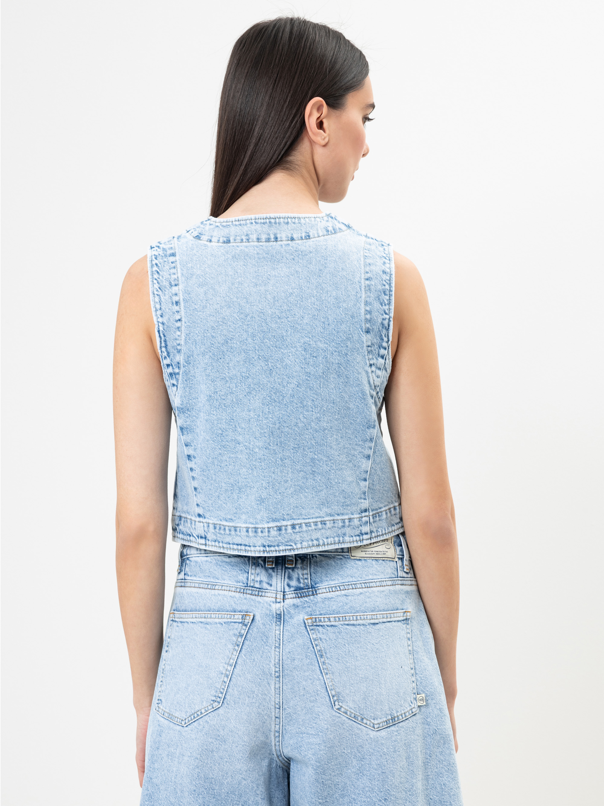 Eine Frau mit langen dunklen Haaren, die eine ärmellose hellblaue Jeansweste und hellblaue Jeans mit hoher Taille trägt, wird von hinten gezeigt und steht vor einem schlichten weißen Hintergrund.