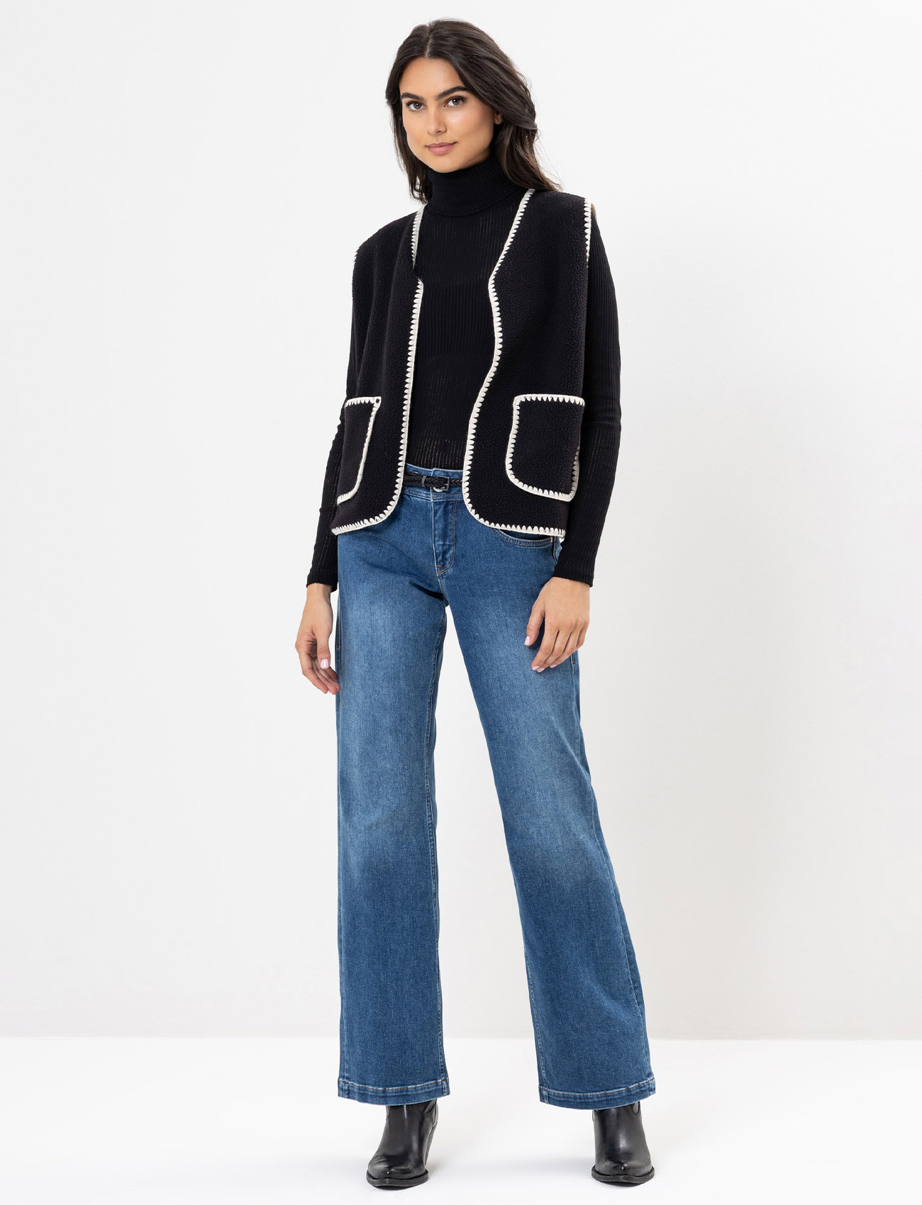 Eine Frau steht vor einem schlichten Hintergrund und trägt einen schwarzen Rollkragenpullover, eine ärmellose schwarze Weste mit weißem Saum, blaue Jeans mit weitem Bein und schwarze Ankle Boots.