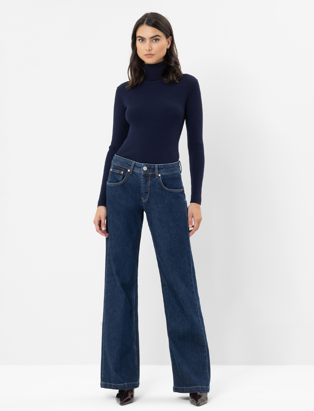 Eine Frau steht vor einem schlichten Hintergrund und trägt einen schwarzen Rollkragenpullover, eine ärmellose schwarze Weste mit weißem Saum, blaue Jeans mit weitem Bein und schwarze Ankle Boots.