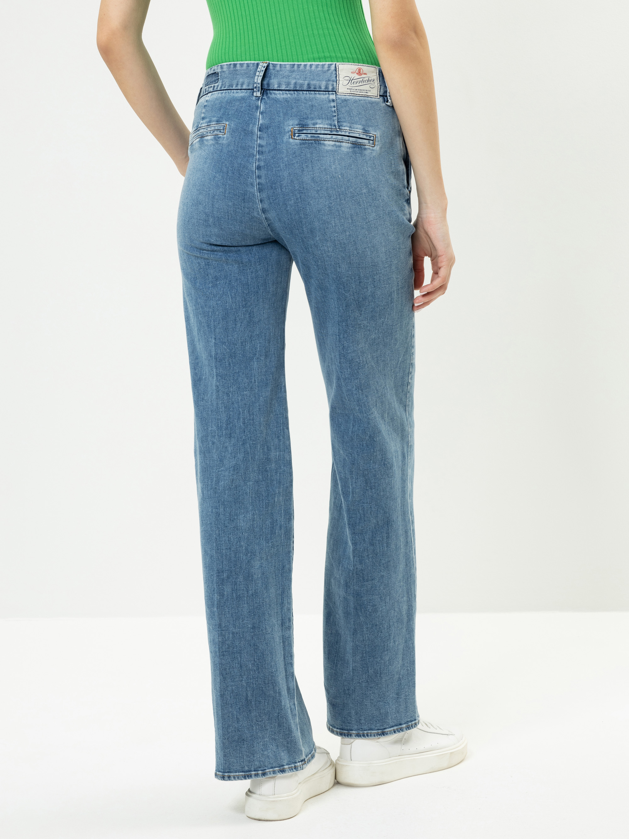 Eine Person, die eine hellblaue Jeans mit geradem Bein, ein grünes geripptes Tanktop und weiße Turnschuhe trägt, wird von hinten vor einem schlichten weißen Hintergrund gezeigt.