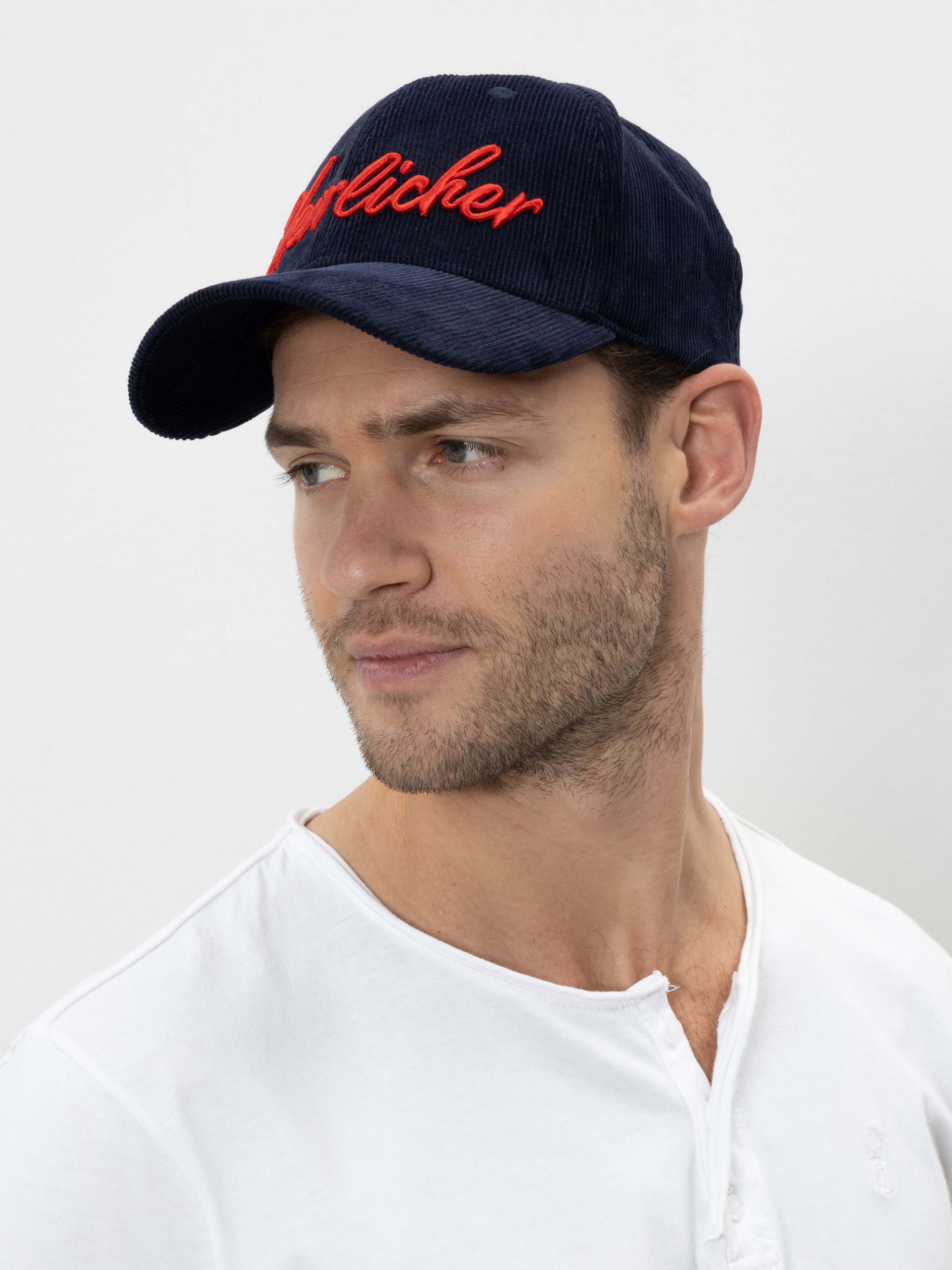 Ein Mann mit heller Haut und kurzem Bart trägt eine marineblaue Mütze mit roter Schreibschrift und ein weißes Hemd mit Knöpfen und blickt leicht zur Seite vor einem schlichten hellen Hintergrund.