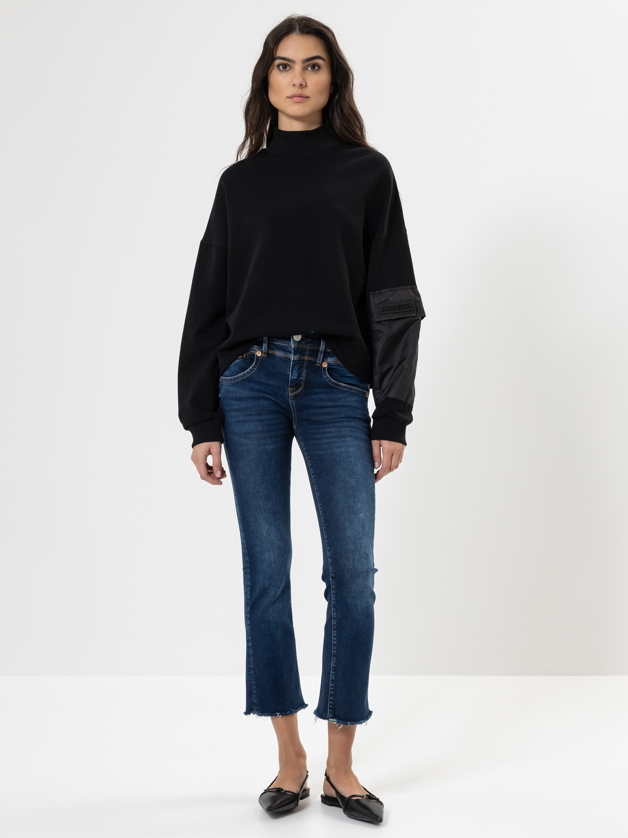 Eine Frau steht vor einem schlichten weißen Hintergrund und trägt ein schwarzes Rollkragen-Sweatshirt, blaue Jeans mit ausgefransten Säumen und schwarze, spitze Schuhe. Sie hat langes gewelltes Haar und einen neutralen Gesichtsausdruck.