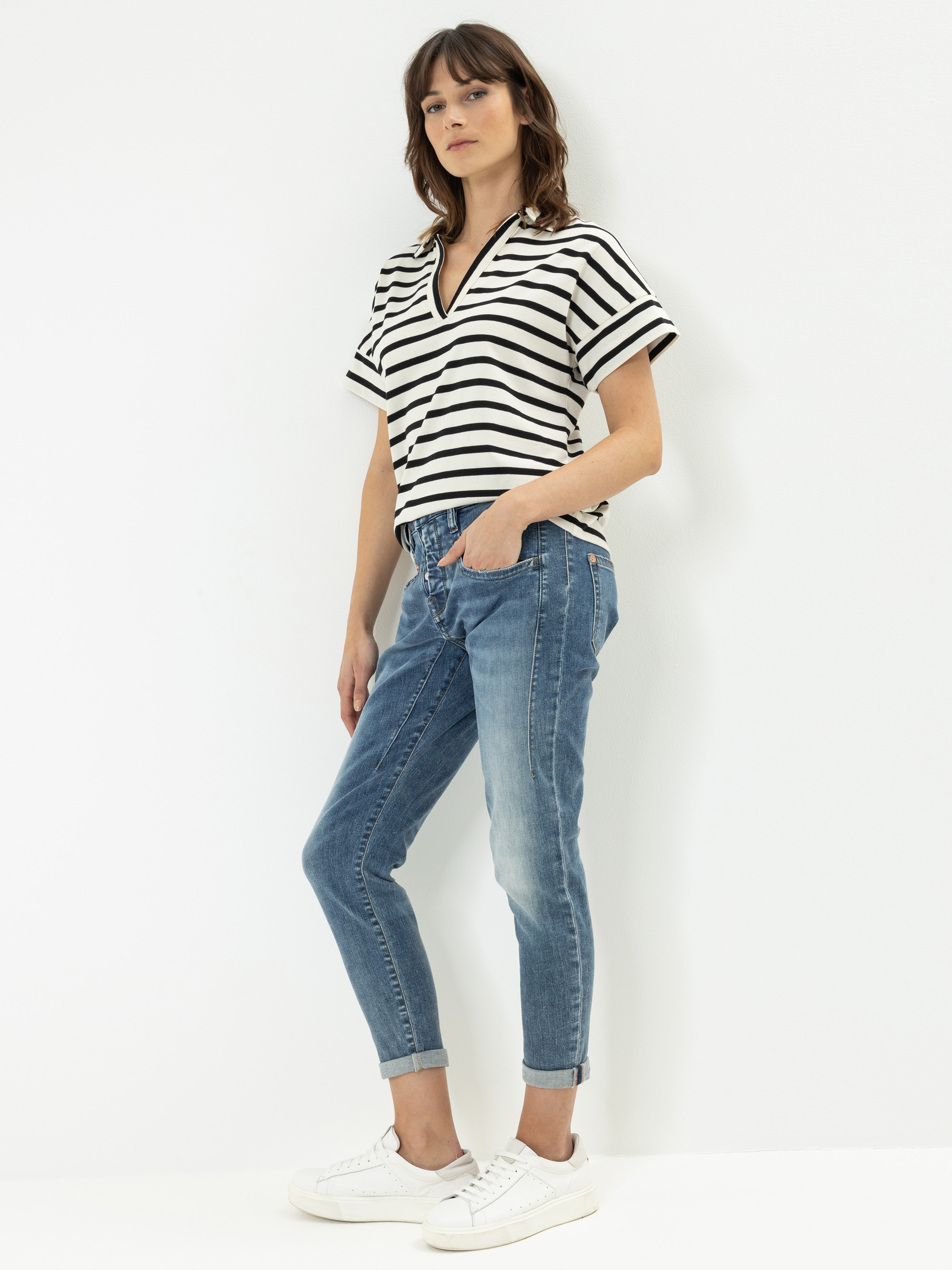 Eine Frau steht vor einem weißen Hintergrund und trägt ein schwarz-weiß gestreiftes kurzärmeliges Oberteil, blaue Jeans mit Bündchen und weiße Turnschuhe. Sie hat glattes braunes Haar und blickt leicht zur Seite.