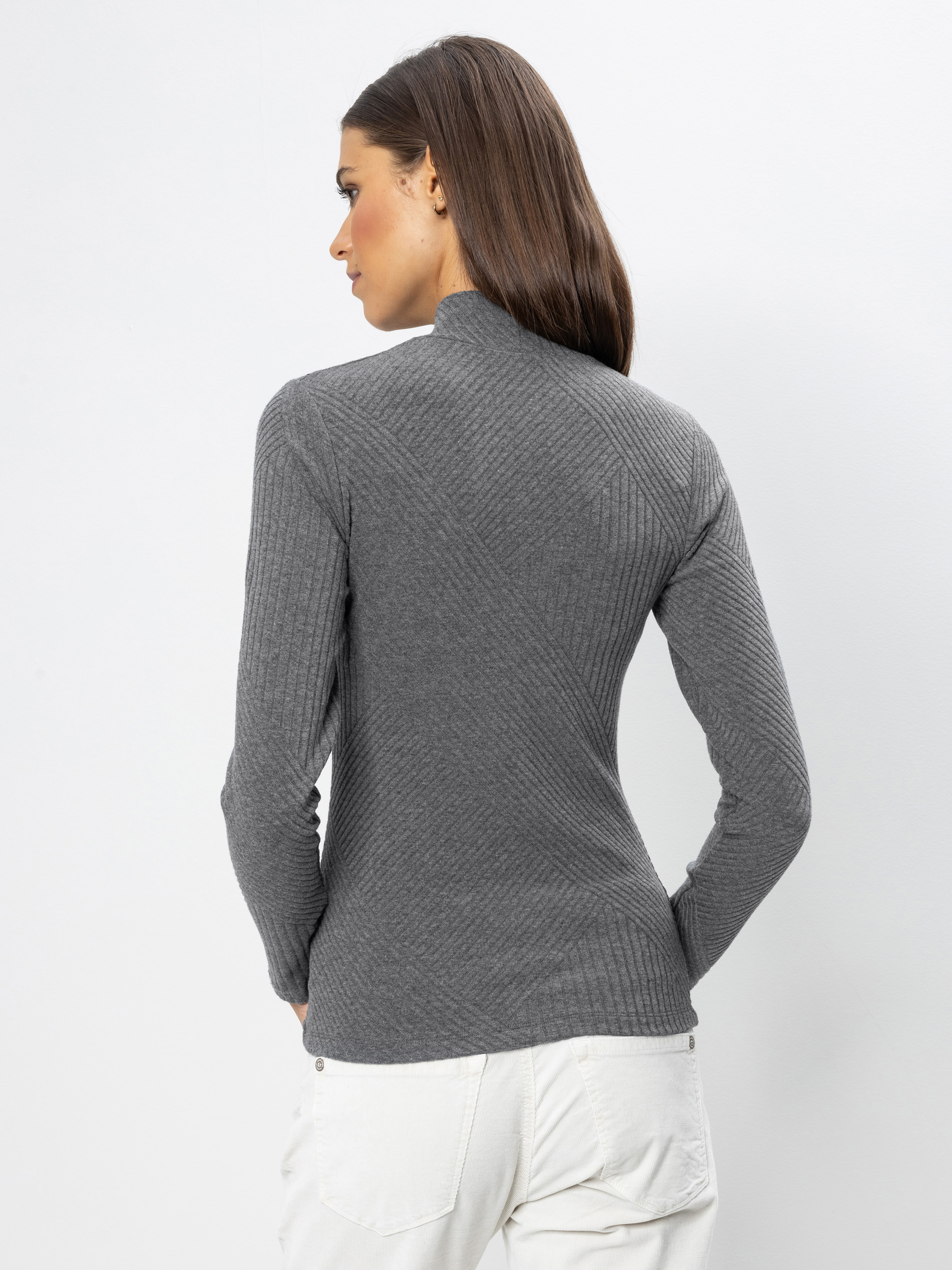 Eine Frau mit glattem braunem Haar ist von hinten zu sehen. Sie trägt ein graues, langärmeliges Oberteil mit Rippenmuster und eine weiße Hose. Sie steht vor einem schlichten weißen Hintergrund.