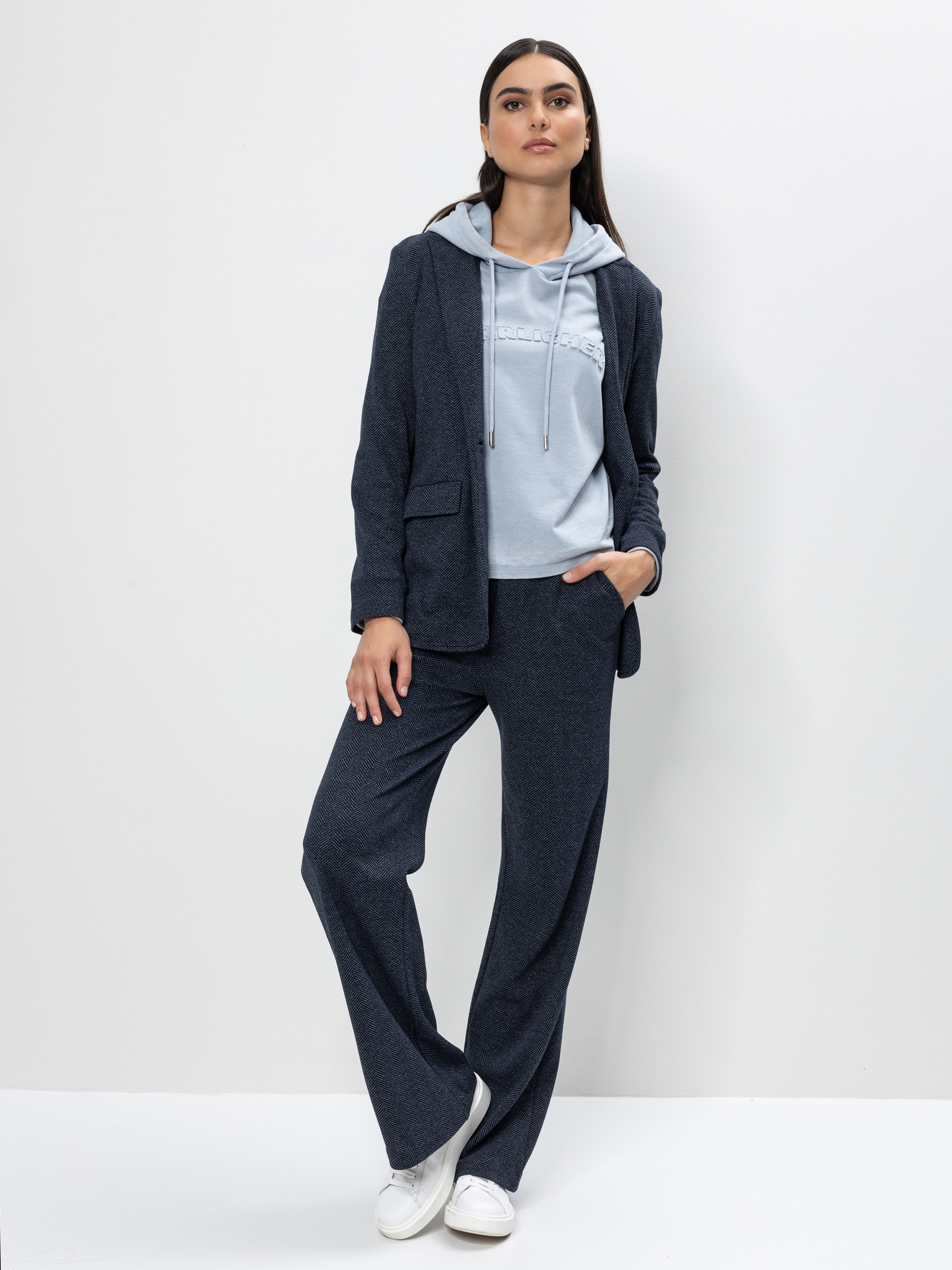 Eine Frau steht vor einer weißen Wand, trägt einen dunkelblauen Anzug mit weiten Hosenbeinen und einen dazu passenden Blazer über einem hellblauen Kapuzenpullover, die linke Hand in der Tasche und weiße Turnschuhe an den Füßen.