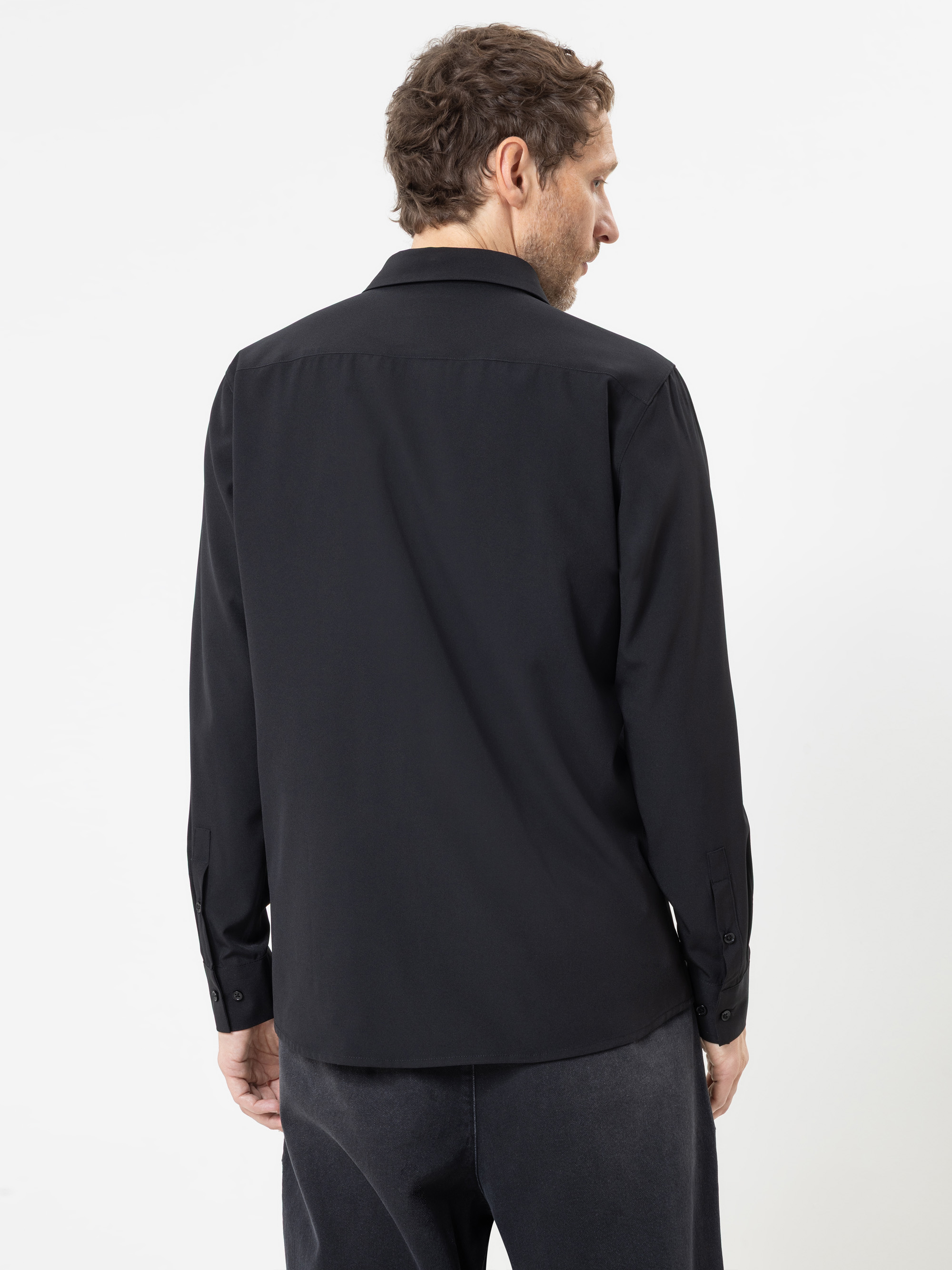 Ein Mann steht mit dem Rücken zur Kamera vor einem schlichten weißen Hintergrund und trägt ein schwarzes Langarmhemd mit Knöpfen sowie eine dunkle Hose.