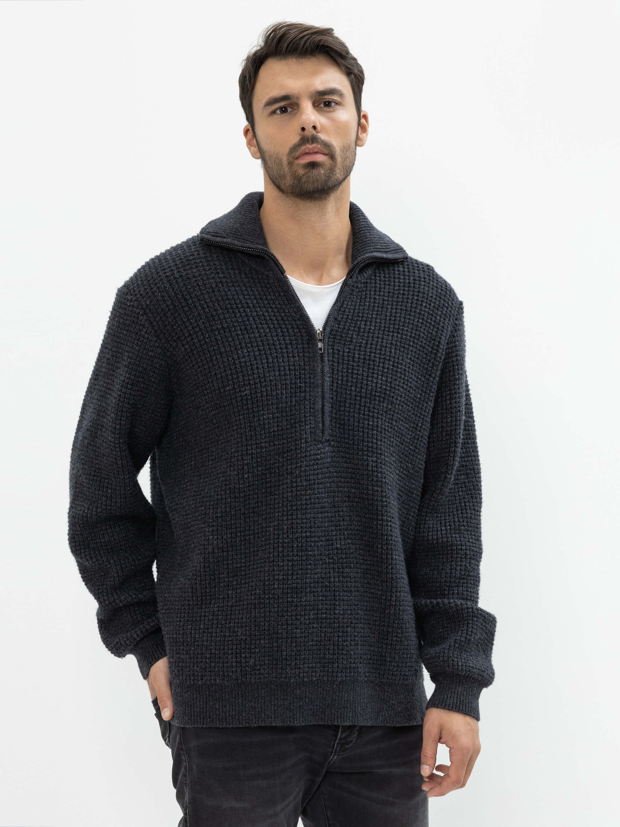 Ein Mann mit dunklem Haar und Bart trägt einen strukturierten, dunklen Pullover mit halbem Reißverschluss über einem weißen Hemd, kombiniert mit einer dunklen Jeans, die vor einem schlichten weißen Hintergrund steht.