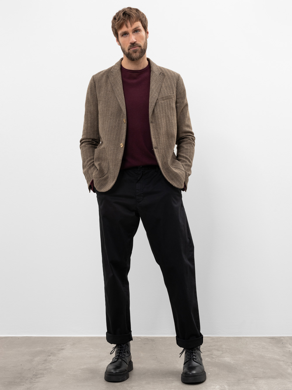 Mann steht vor einer weißen Wand, trägt einen hellbraunen Blazer, einen weinroten Pullover, eine schwarze Hose und schwarze Schnürstiefel; Hände in den Taschen, schaut mit neutralem Blick in die Kamera.