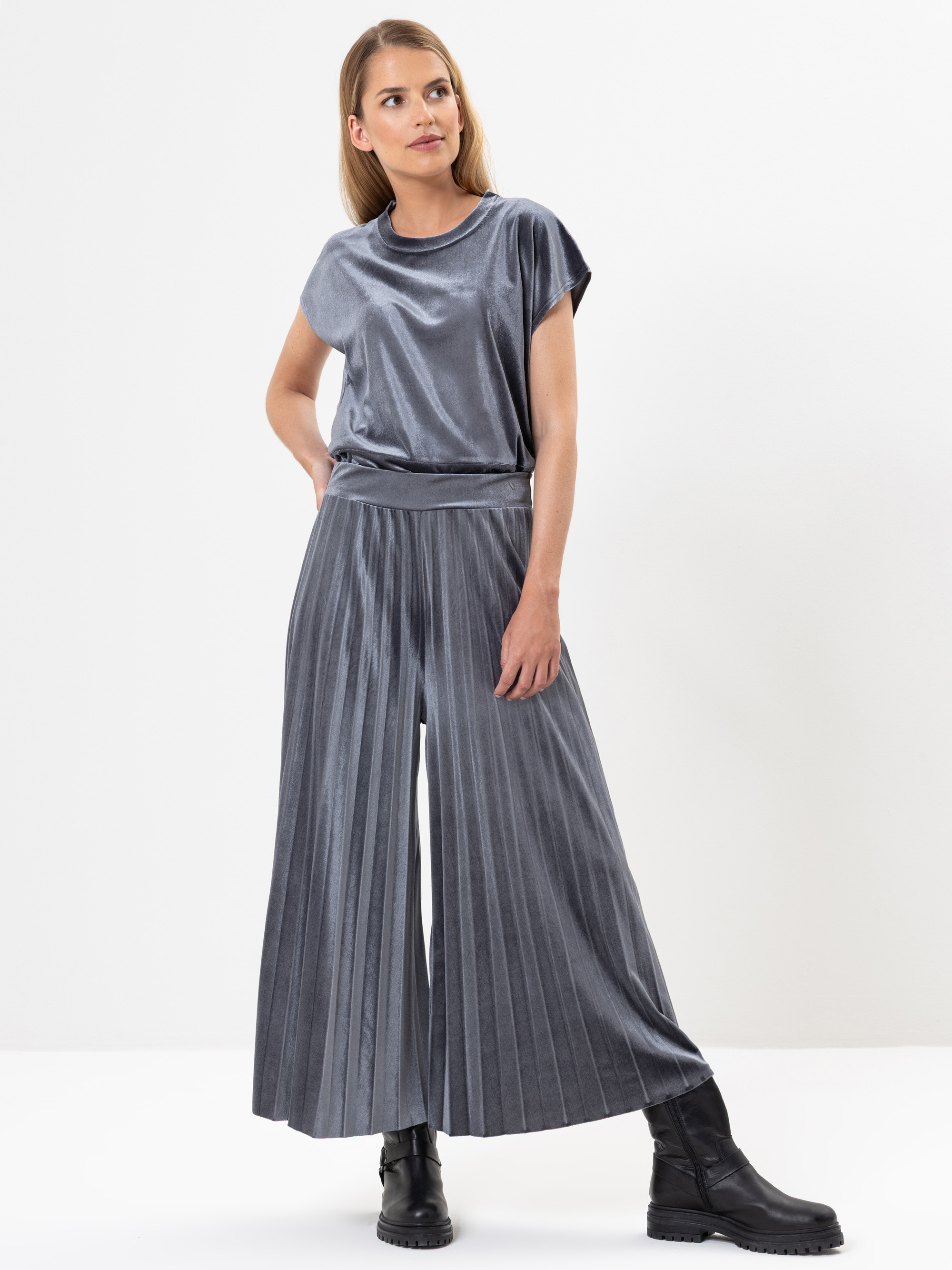 Eine Frau steht in einem Studio und trägt ein silbern glänzendes, kurzärmeliges Oberteil und eine dazu passende, weit geschnittene Hose mit Falten. Sie kombiniert das Outfit mit schwarzen Ankle Boots und hat glattes, schulterlanges blondes Haar.