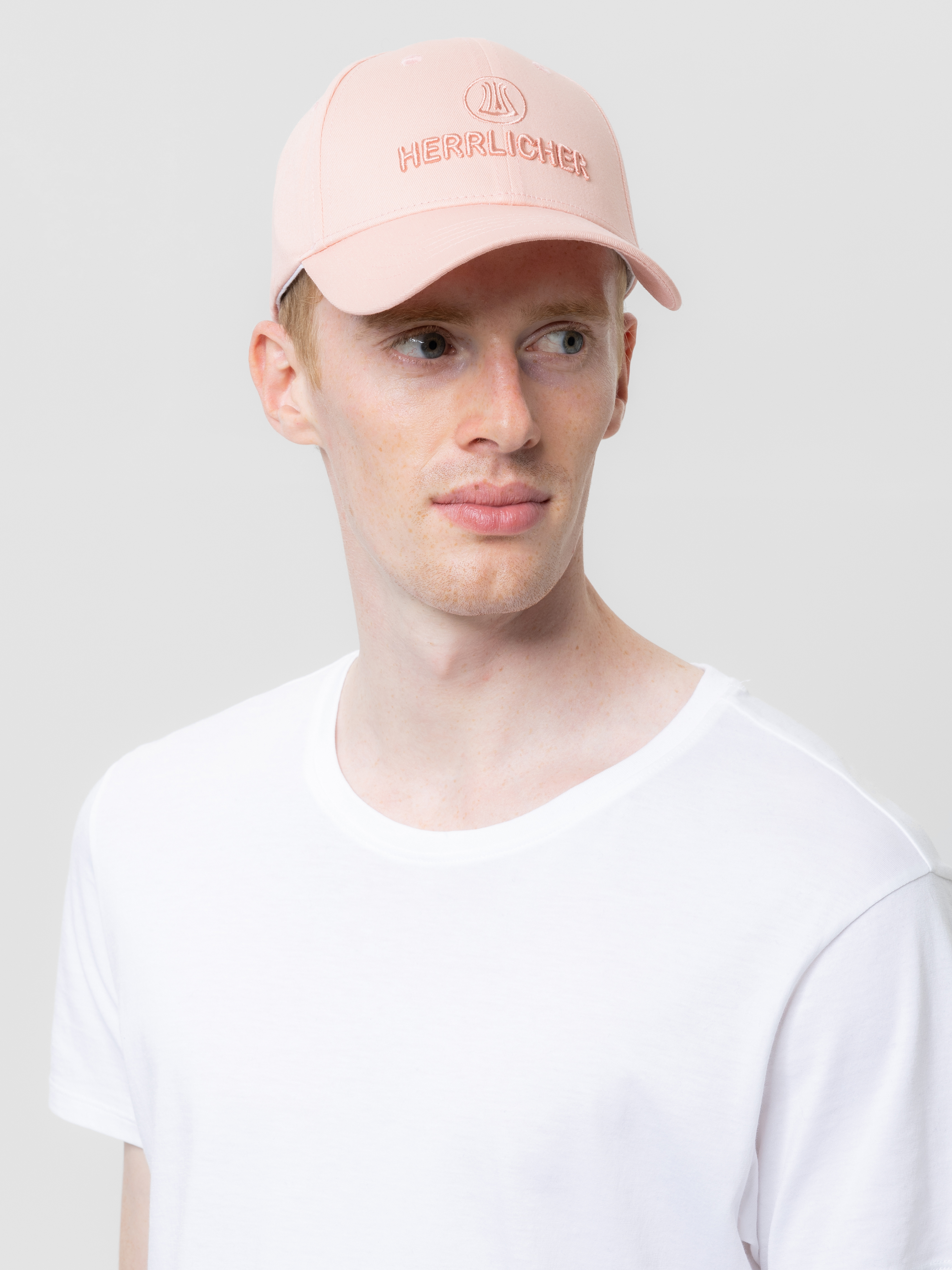 Ein junger Mann, der eine hellrosa Baseballkappe mit HERRLICHER-Stickerei und ein schlichtes weißes T-Shirt trägt und vor einem hellgrauen Hintergrund leicht nach rechts schaut.