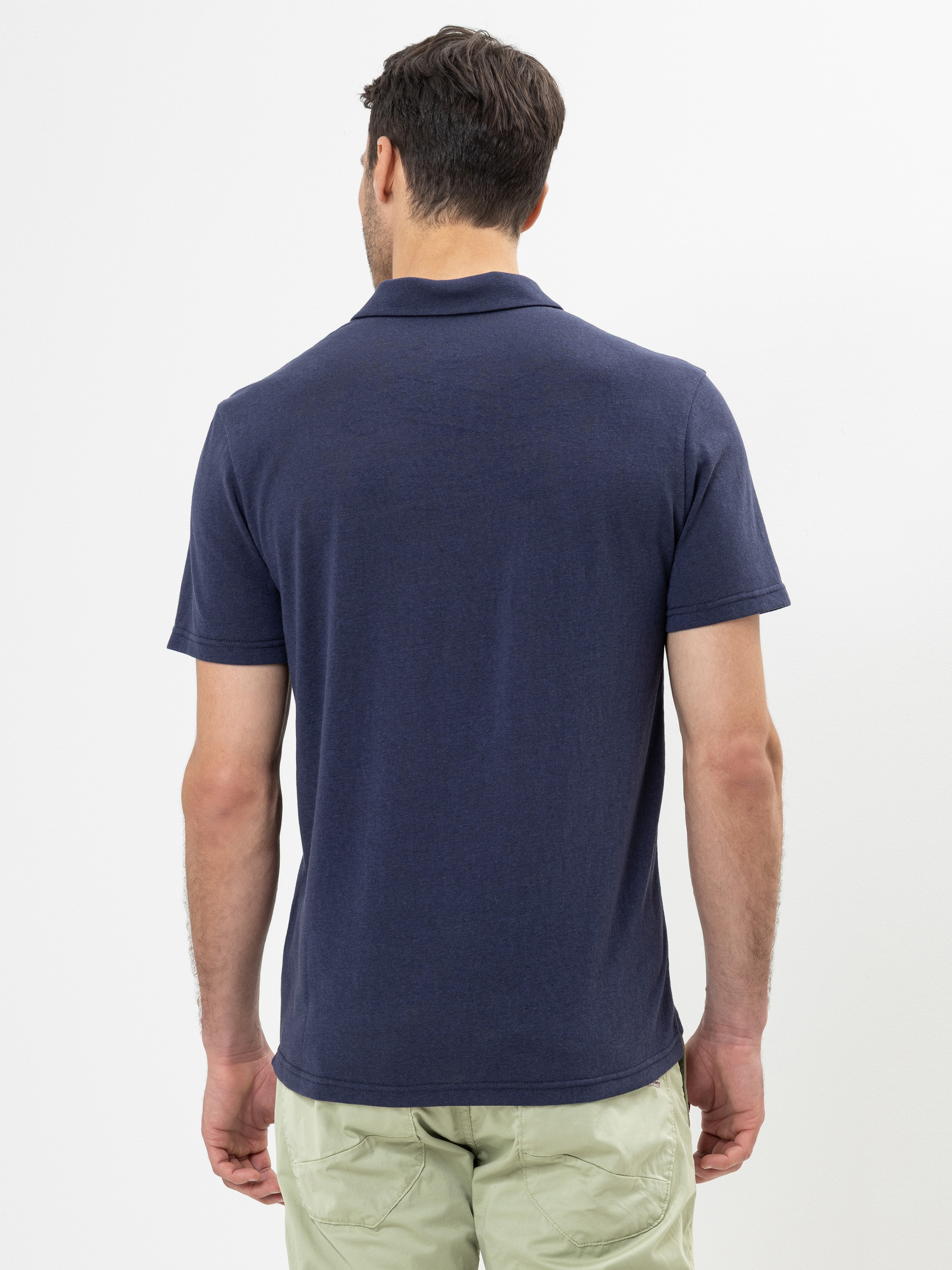 Ein Mann steht von der Kamera abgewandt in einem kurzärmeligen marineblauen Polohemd und einer hellen Hose vor einem schlichten weißen Hintergrund.