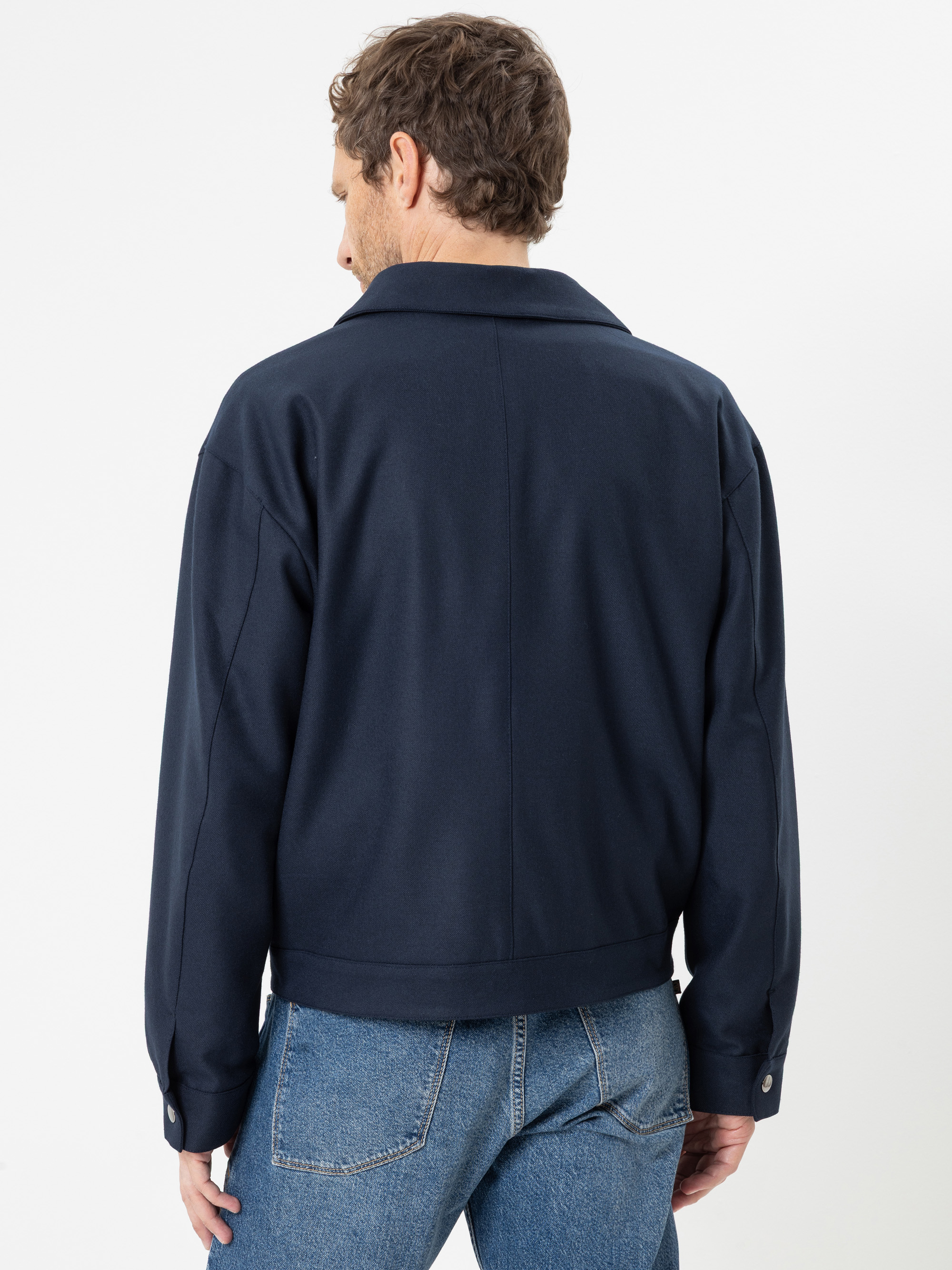 Eine Person, die eine dunkelblaue Jacke und blaue Jeans trägt, ist von hinten vor einem schlichten weißen Hintergrund zu sehen.