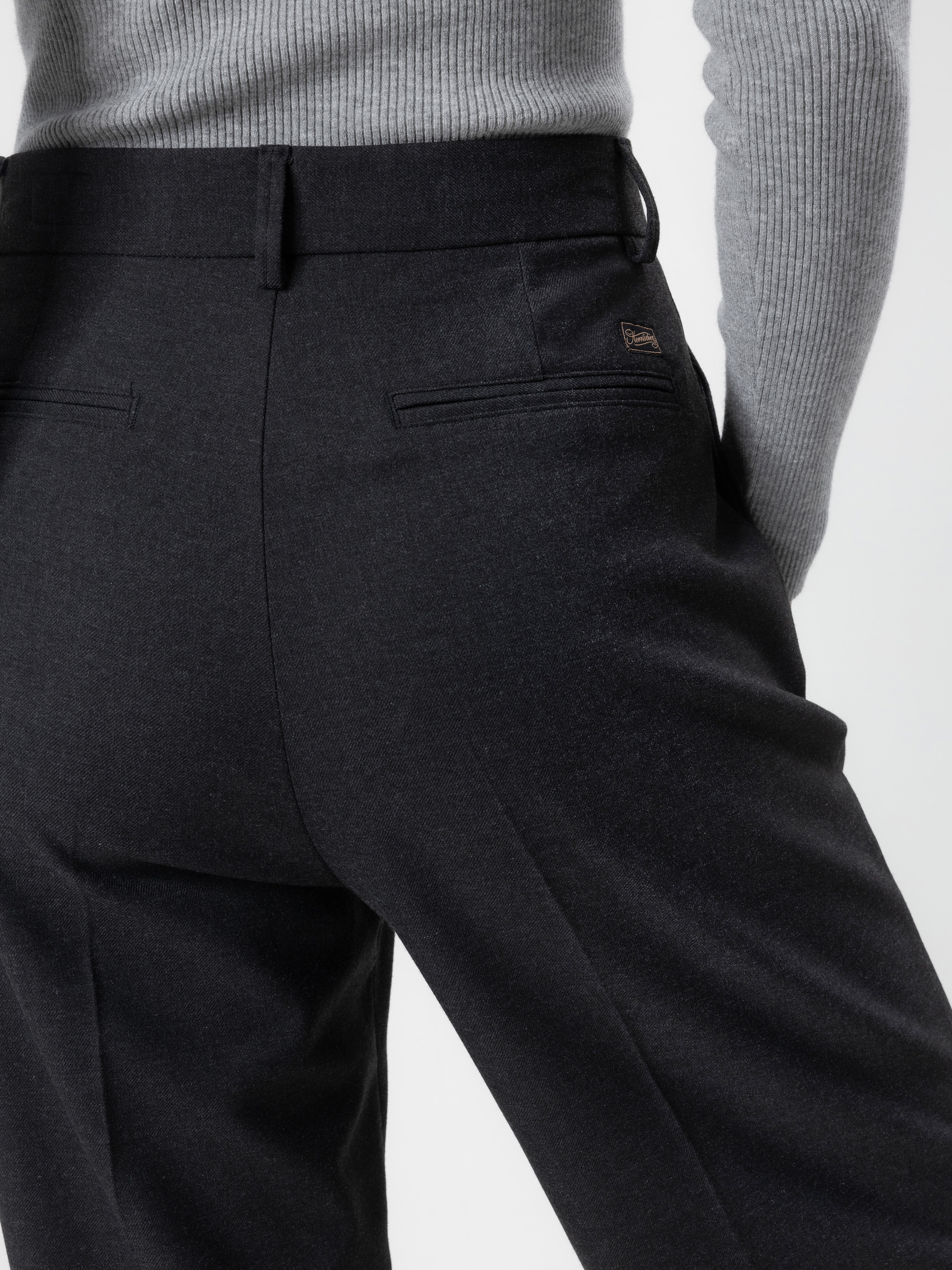 Eine Person, die eine hochgeschnittene, dunkle, maßgeschneiderte Hose und einen hellgrauen, gerippten Pullover trägt, steht mit den Händen in den Taschen, von hinten und von der Taille abwärts gezeigt.