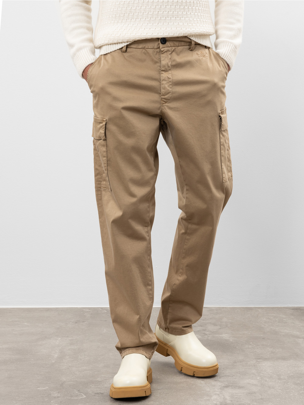 Eine Person in hellbraunen Cargohosen, einem cremefarbenen Strickpullover und weißen Stiefeln steht mit den Händen in den Taschen vor einem schlichten Hintergrund. Ihr Oberkörper ist teilweise außerhalb des Bildausschnitts.