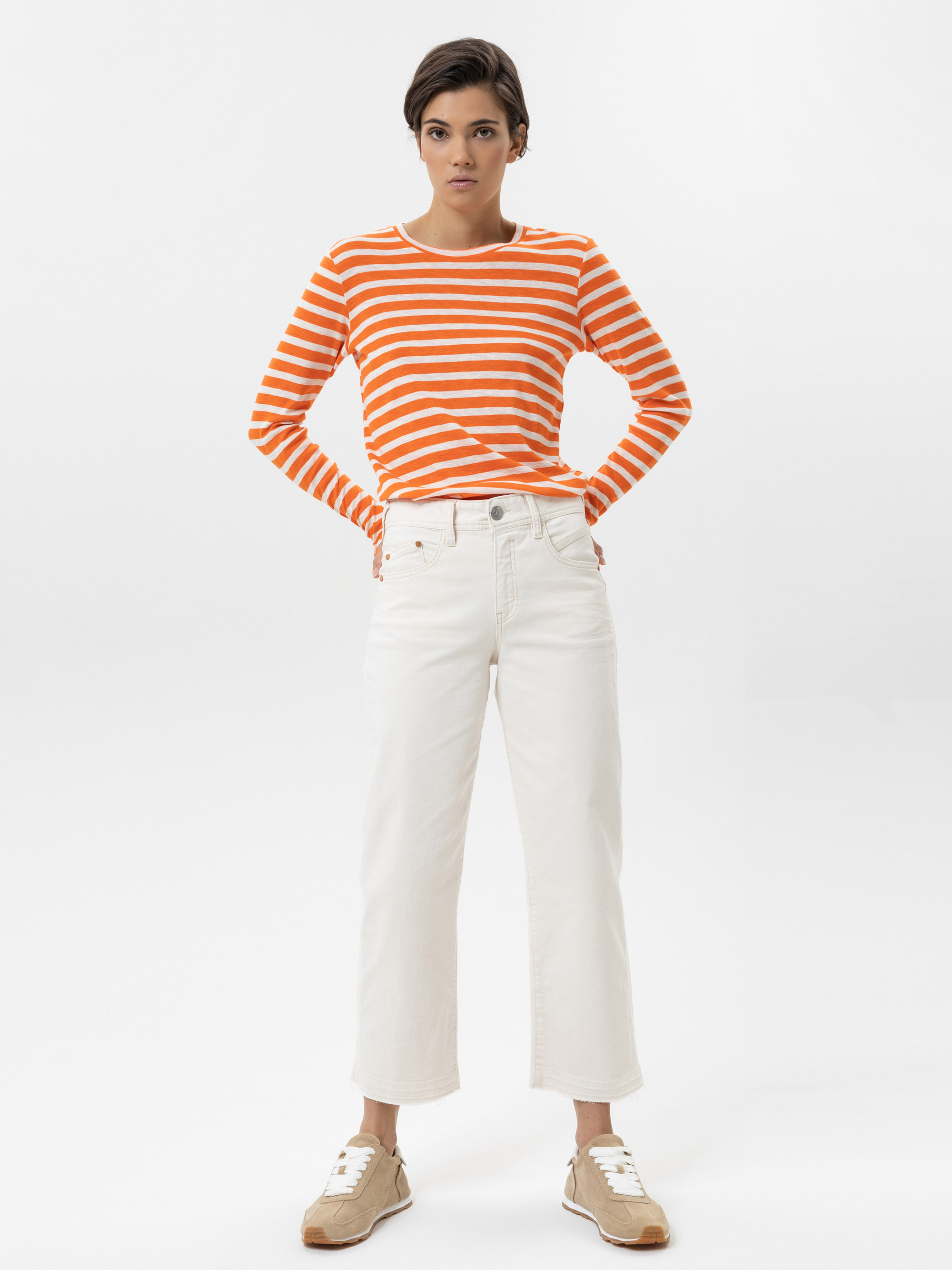Eine Person steht mit dem Gesicht nach vorne, trägt ein orange-weiß gestreiftes langärmeliges Hemd, weiße Jeans mit hoher Taille und beigefarbene Turnschuhe vor einem schlichten weißen Hintergrund.