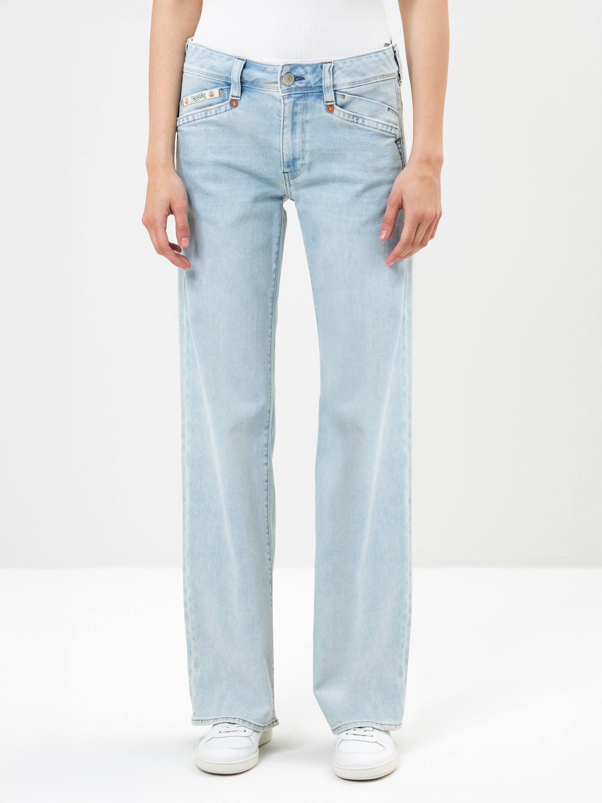 Eine Person, die eine hellblaue Jeans mit hoher Taille und geradem Bein und mehreren Taschen auf der Vorderseite trägt, kombiniert mit einem weißen Oberteil und weißen Turnschuhen, steht vor einem schlichten weißen Hintergrund.