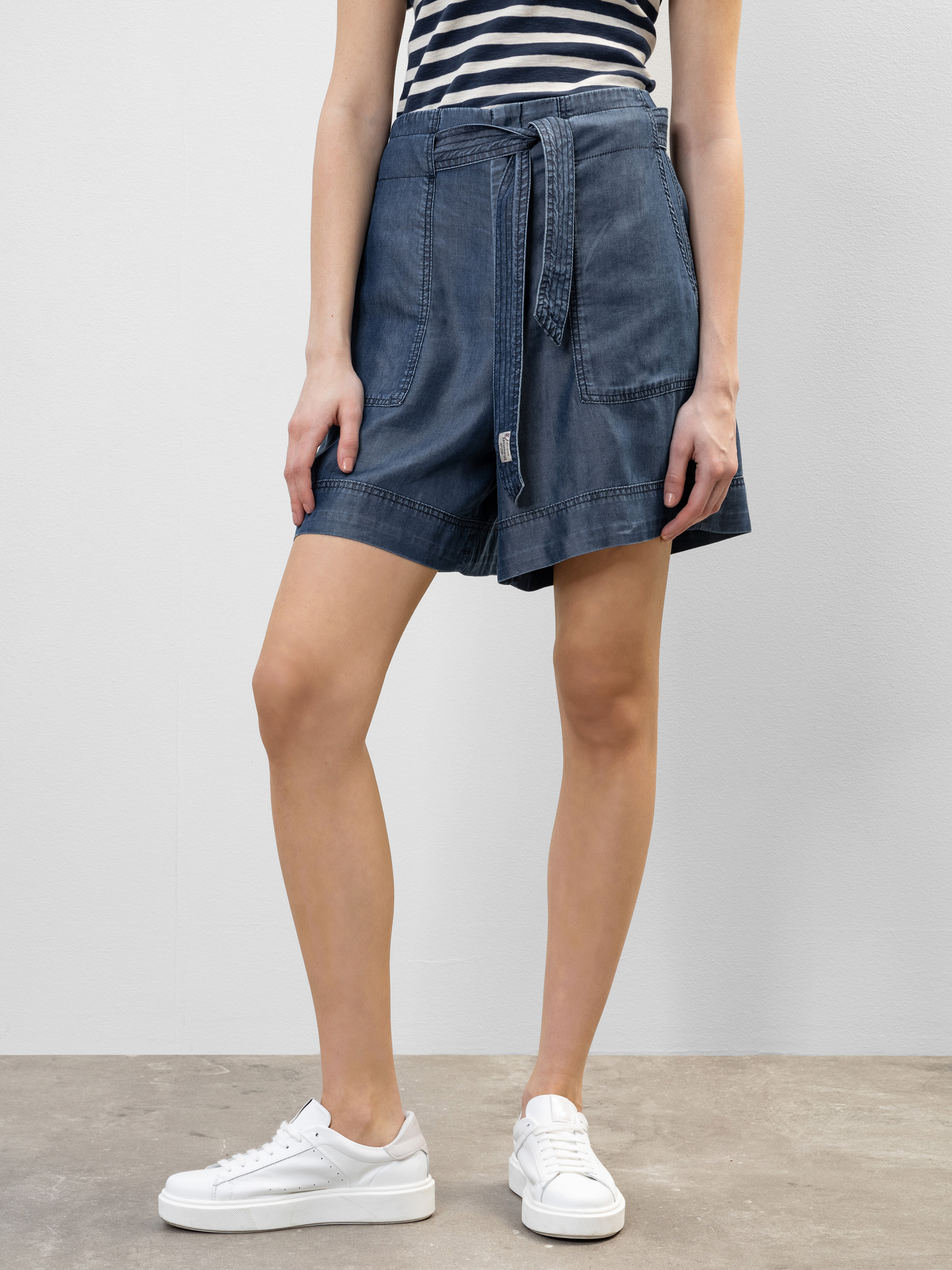 Eine Person, die ein schwarz-weiß gestreiftes ärmelloses Oberteil, lockere Jeansshorts mit passendem Gürtel und weiße Turnschuhe trägt, steht vor einer schlichten hellen Wand.