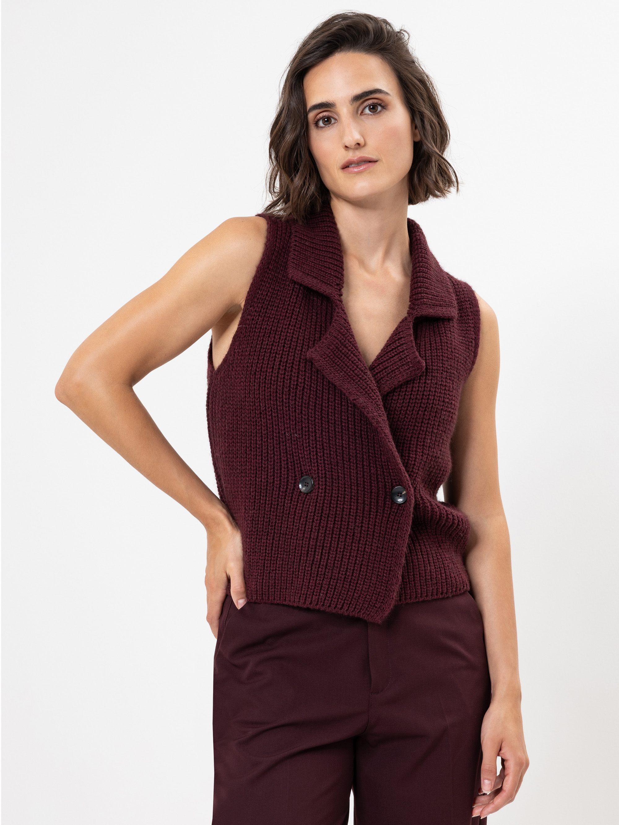 Eine Frau mit kurzen braunen Haaren trägt eine ärmellose, weinrote Strickweste mit Knöpfen über einer passenden weinroten Hose und steht vor einem schlichten weißen Hintergrund.