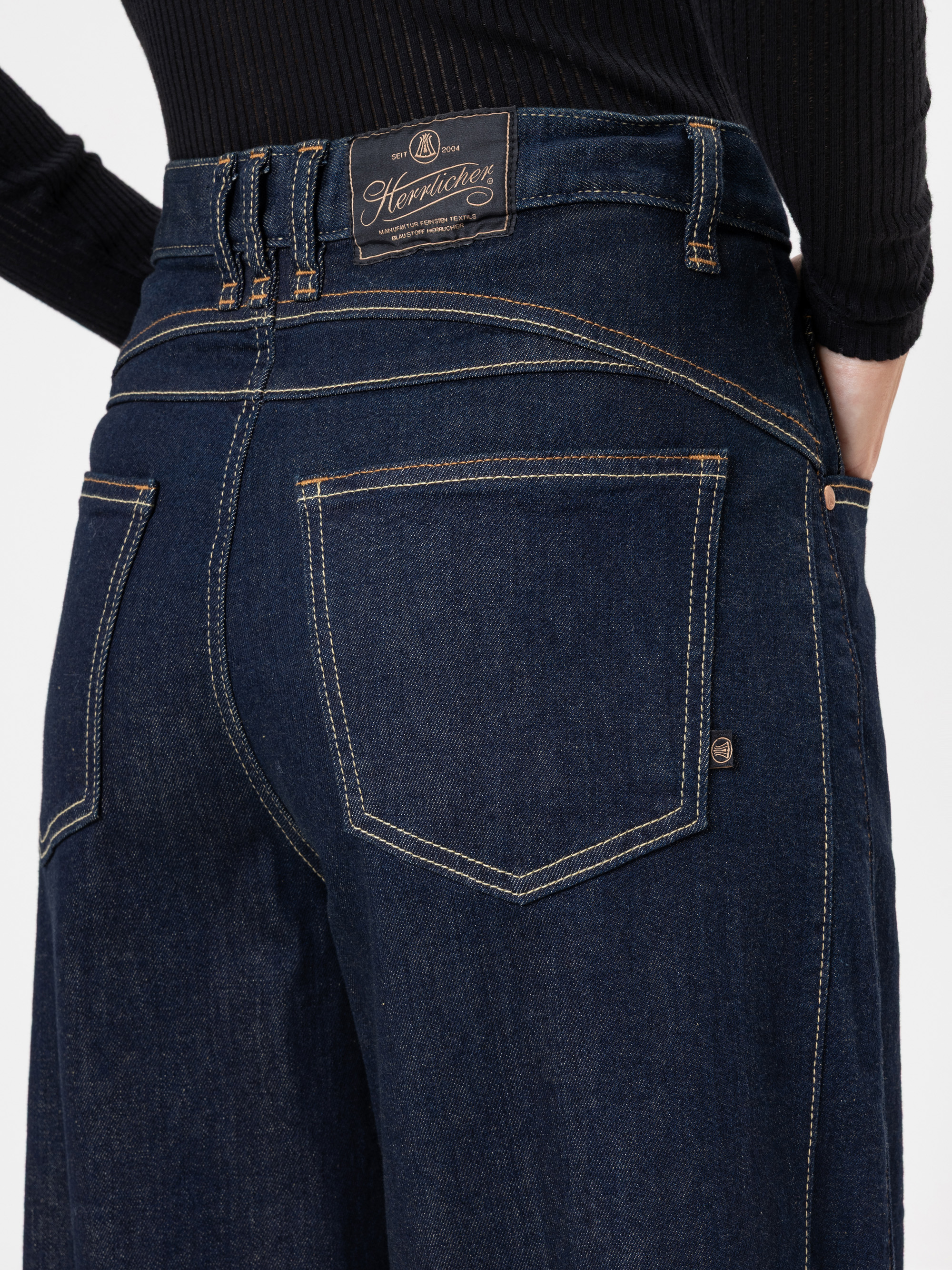 Nahaufnahme einer Person, die eine dunkelblaue Jeans mit hoher Taille und Kontrastnähten sowie ein schwarzes geripptes Oberteil trägt. Die Jeans hat große Gesäßtaschen und ein sichtbares Markenetikett am Bund.