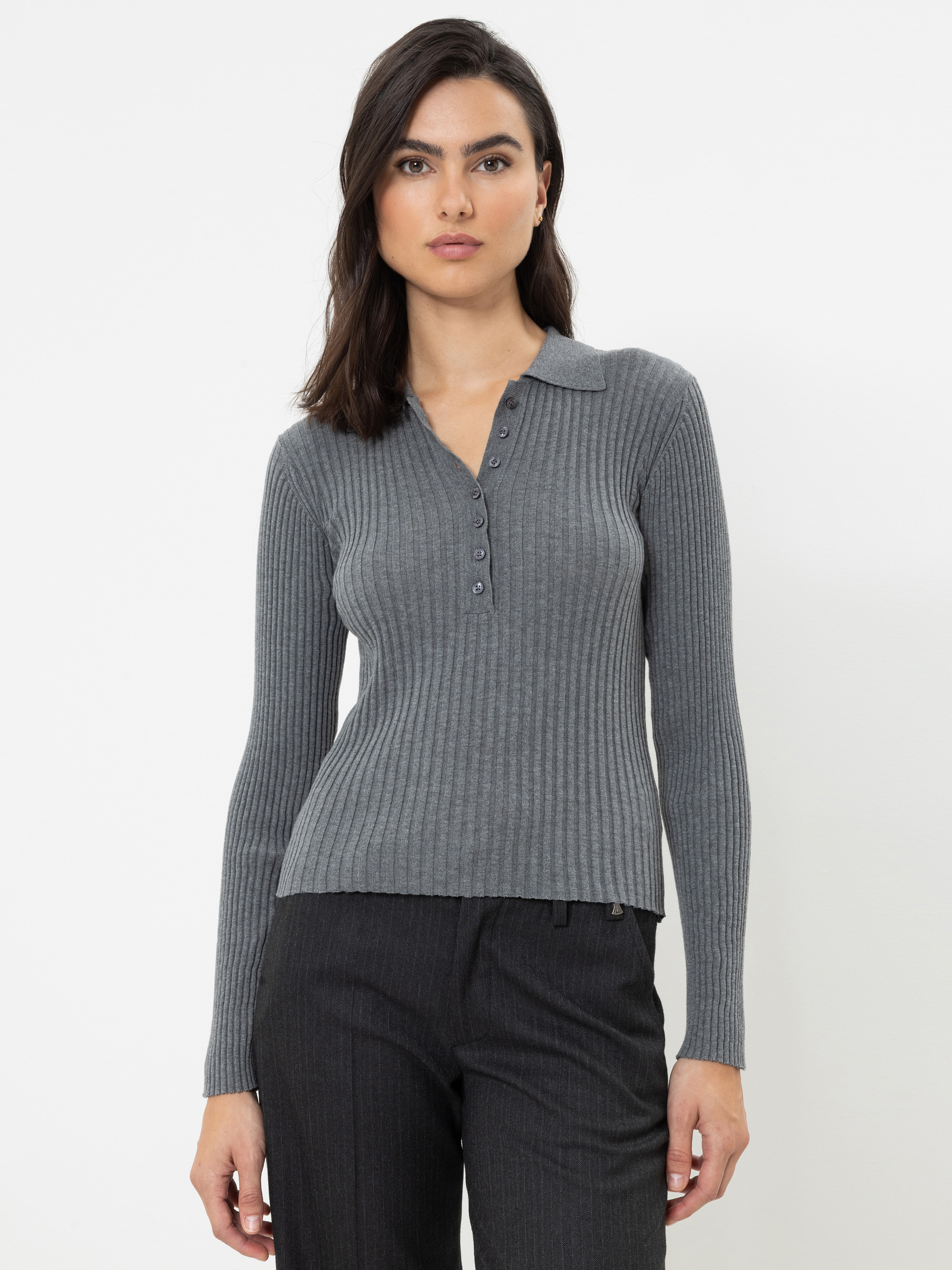 Eine Frau mit glattem braunem Haar trägt ein eng anliegendes, geripptes, graues Henley-Shirt mit langen Ärmeln und Knöpfen sowie eine dunkle Hose und steht vor einem schlichten weißen Hintergrund.