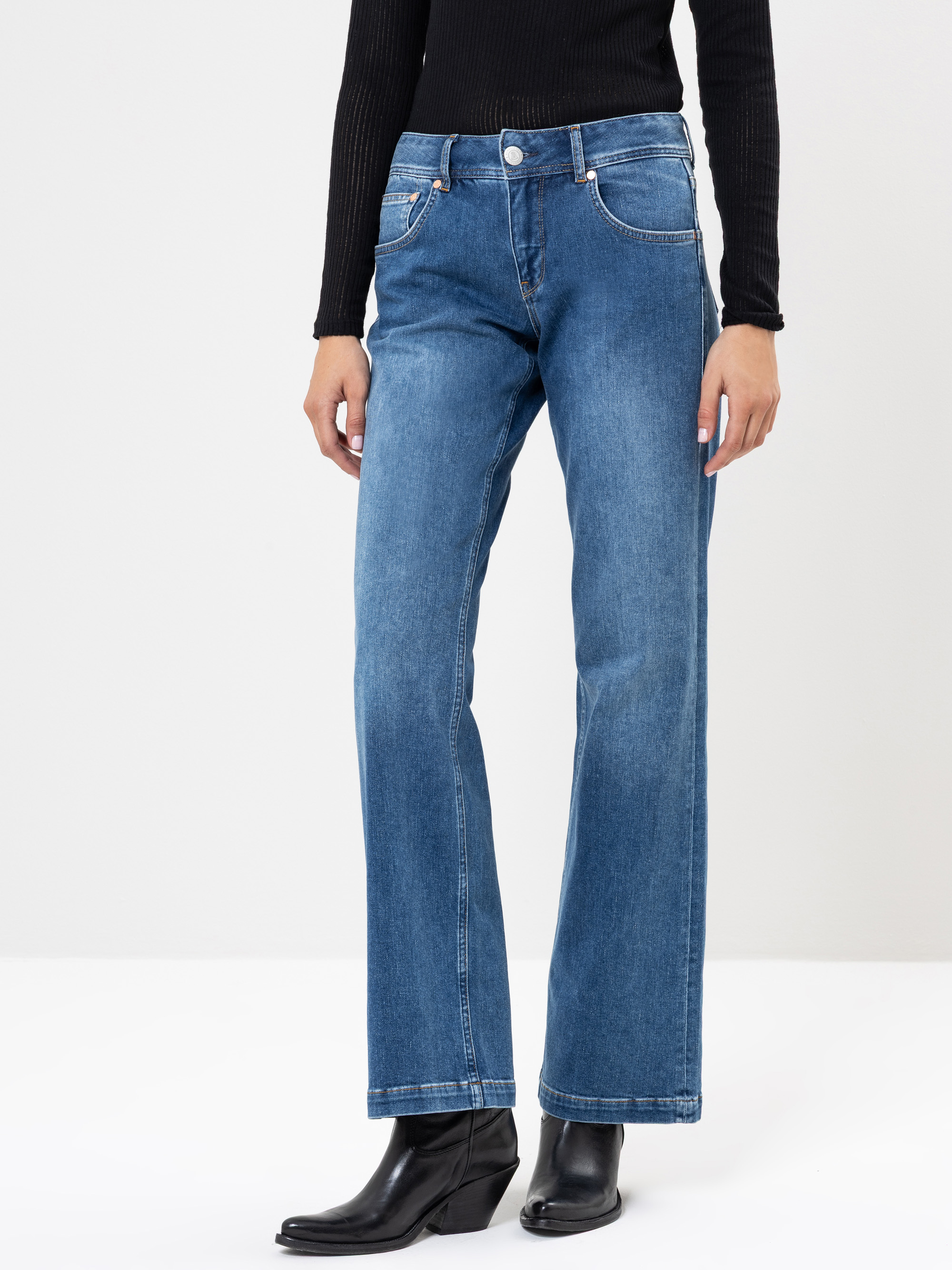 Eine Person, die blaue Jeans mit geradem Bein, ein schwarzes geripptes langärmeliges Oberteil und schwarze Stiefeletten trägt, steht vor einem schlichten hellen Hintergrund.