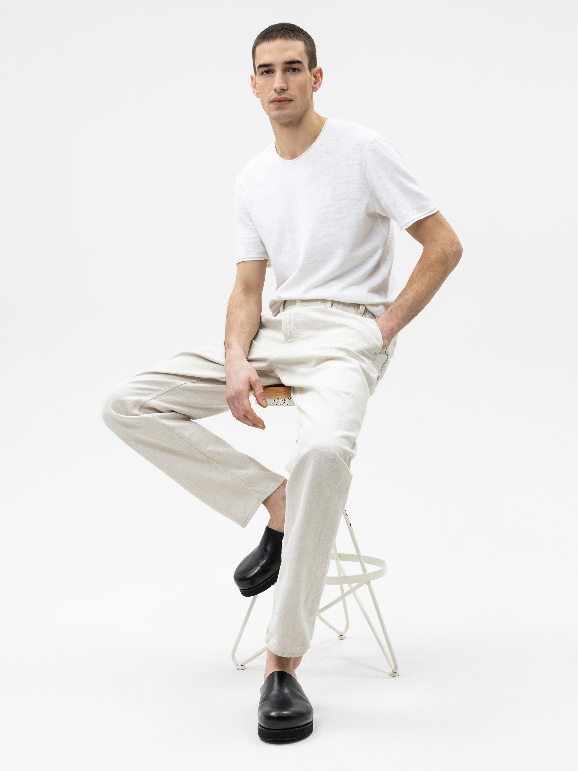 Ein Mann in einem weißen T-Shirt und einer hellbeigen Hose sitzt auf einem weißen Klappstuhl vor einem schlichten weißen Hintergrund. Er trägt schwarze Schuhe und hat kurze dunkle Haare. Er sieht entspannt aus und hat eine Hand in der Tasche.