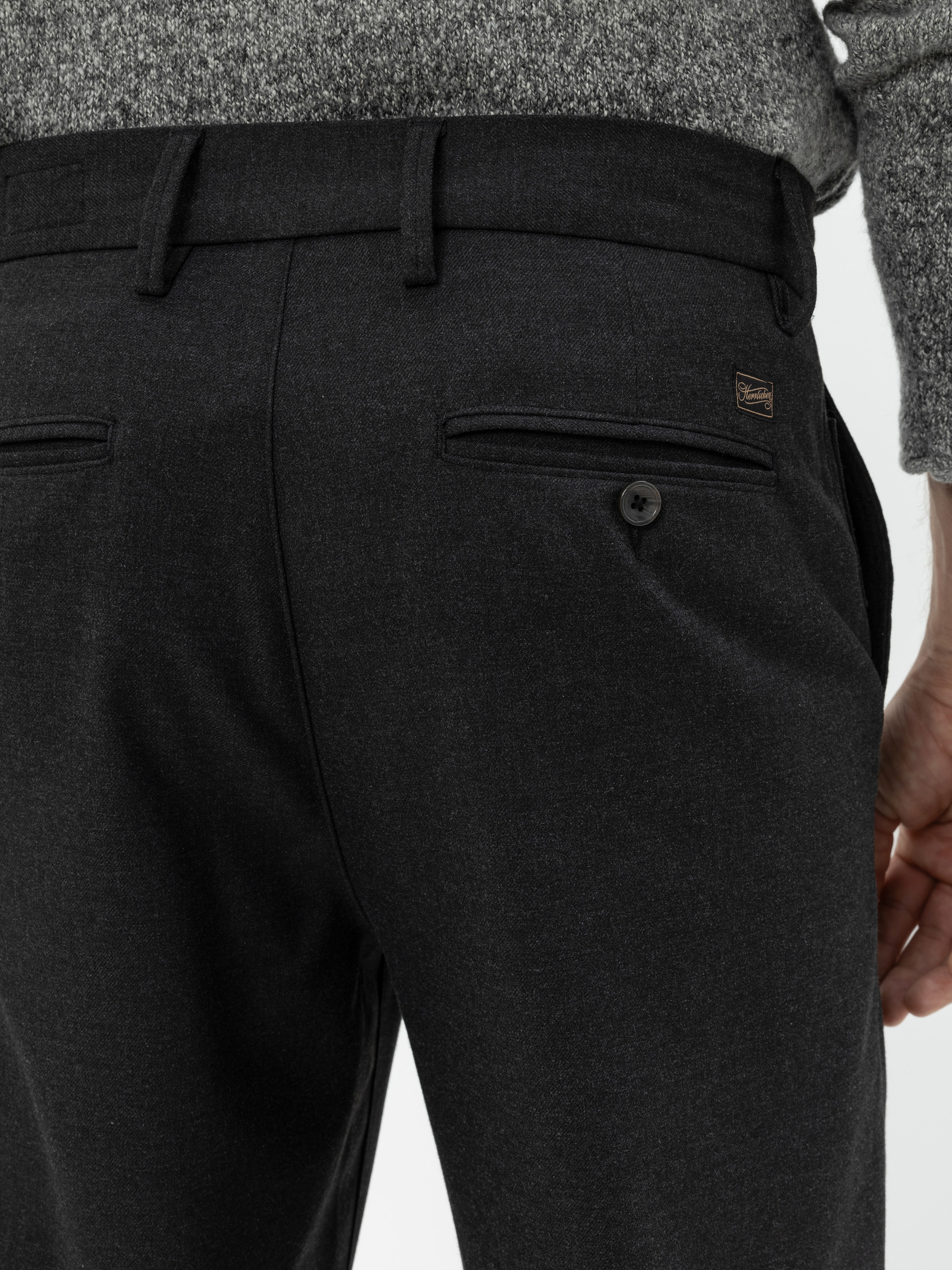 Nahaufnahme einer Person, die eine dunkelgraue Anzughose mit geknöpften Gesäßtaschen und einen grauen Strickpullover trägt, wobei die Taille und der obere Beinbereich von hinten gezeigt werden.