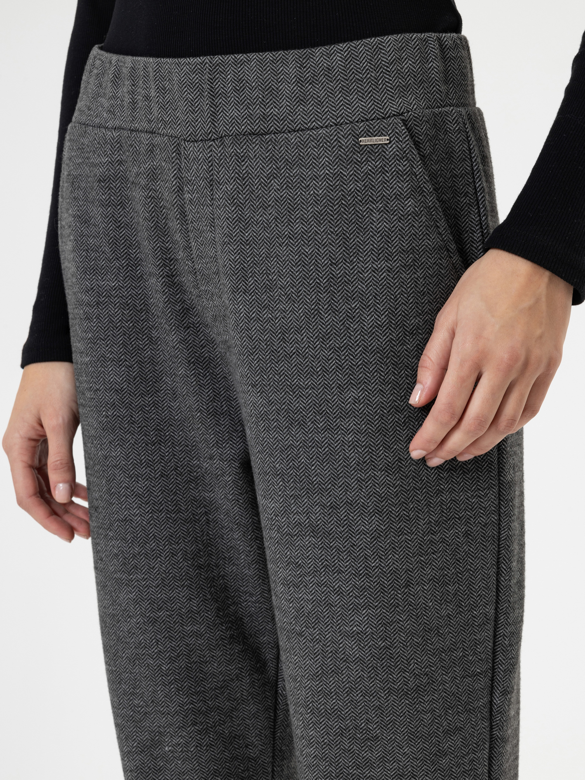Eine Person, die eine graue Hose mit Fischgrätenmuster und ein langärmeliges schwarzes Oberteil trägt, wobei eine Hand an der Seite ruht und die andere teilweise sichtbar ist.