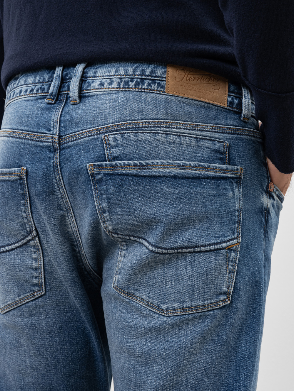 Nahaufnahme des Rückens einer Person, die eine blaue Jeans mit großen Gesäßtaschen und einem braunen Lederaufnäher am Bund trägt, kombiniert mit einem dunklen Pullover.