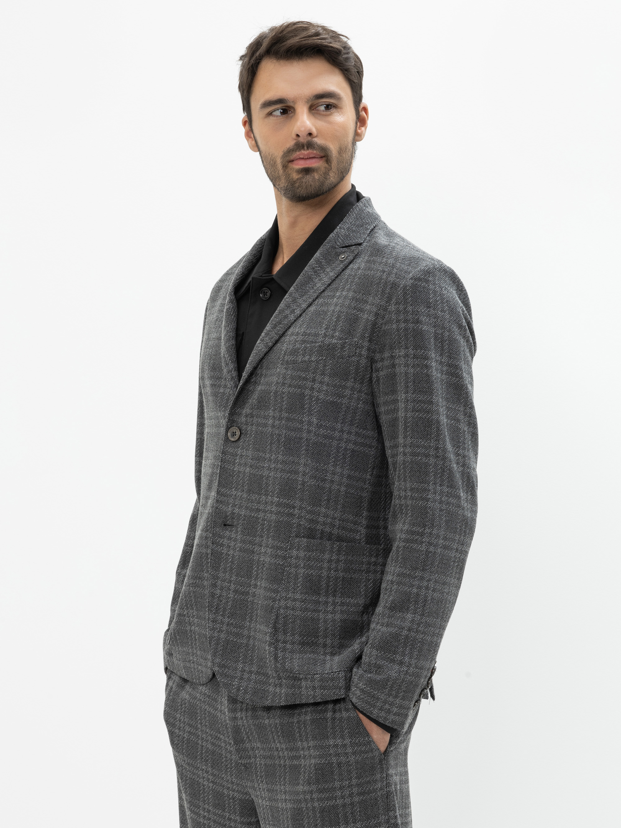 Ein Mann mit kurzen dunklen Haaren und einem Bart trägt einen grau karierten Anzug mit einem schwarzen Hemd. Er hat die Hände in den Taschen und blickt vor einem schlichten weißen Hintergrund zur Seite.