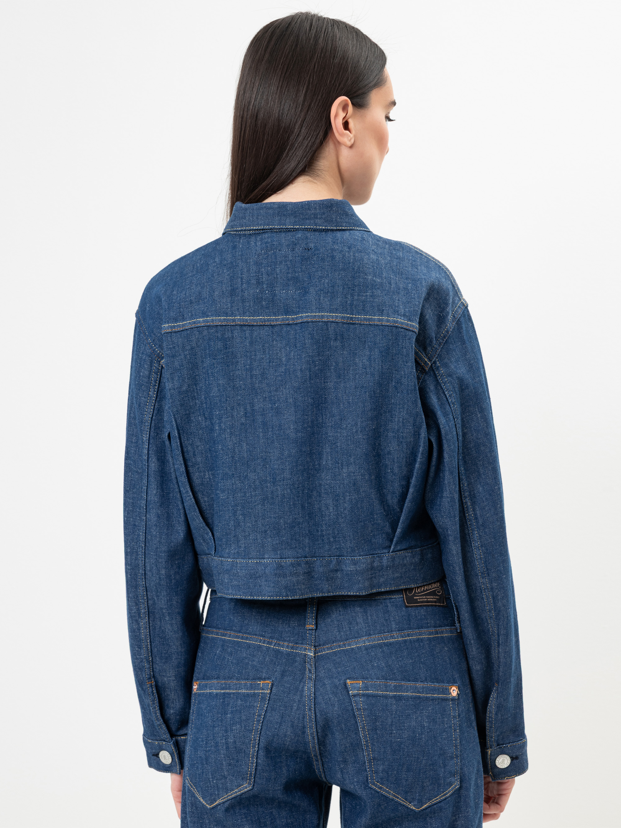 Eine Frau mit langen dunklen Haaren, von hinten gesehen, trägt eine blaue Jeansjacke und dazu passende Jeans mit hoher Taille vor einem schlichten weißen Hintergrund.