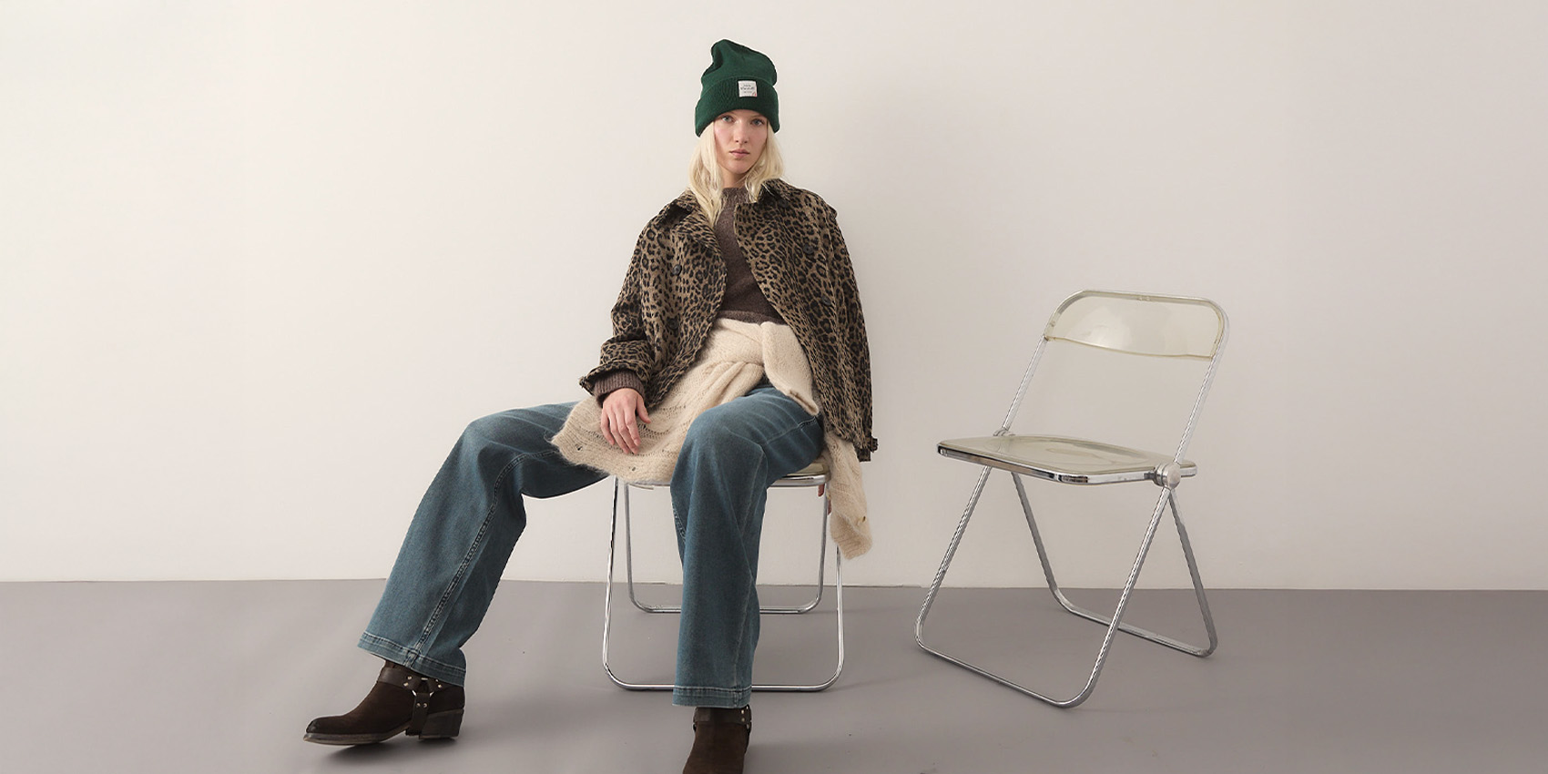 Eine Person, die eine grüne Mütze, eine Jacke mit Leopardenmuster, Jeans mit weitem Bein und braune Stiefel trägt, sitzt auf einem Metallstuhl neben einem leeren transparenten Klappstuhl vor einem unifarbenen Hintergrund.