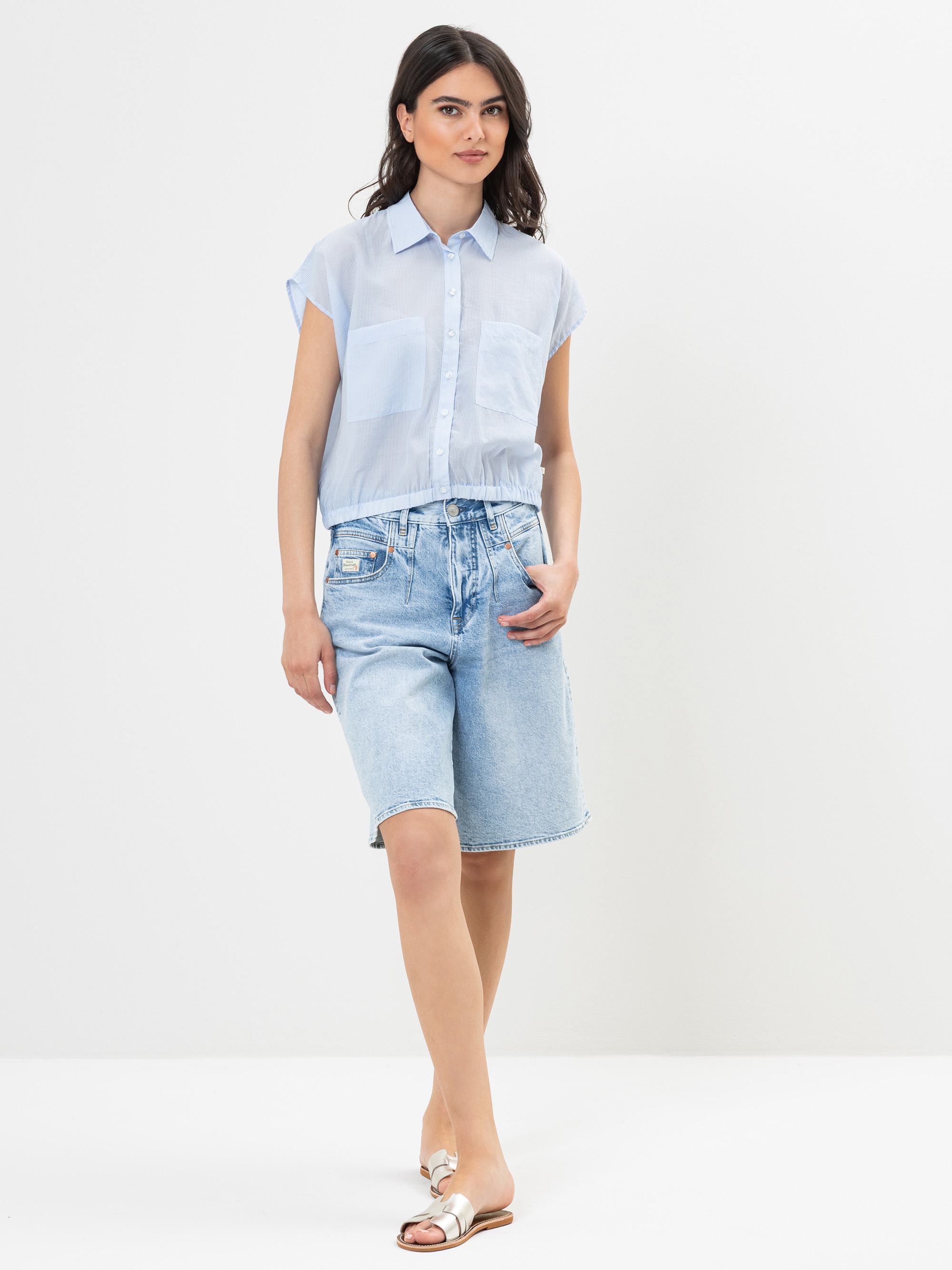 Eine Frau steht vor einem schlichten weißen Hintergrund und trägt ein hellblaues, kurzärmeliges Button-up-Hemd, weite, knielange Jeans-Shorts und weiße Sandalen. Sie hat langes dunkles Haar und einen entspannten Gesichtsausdruck.