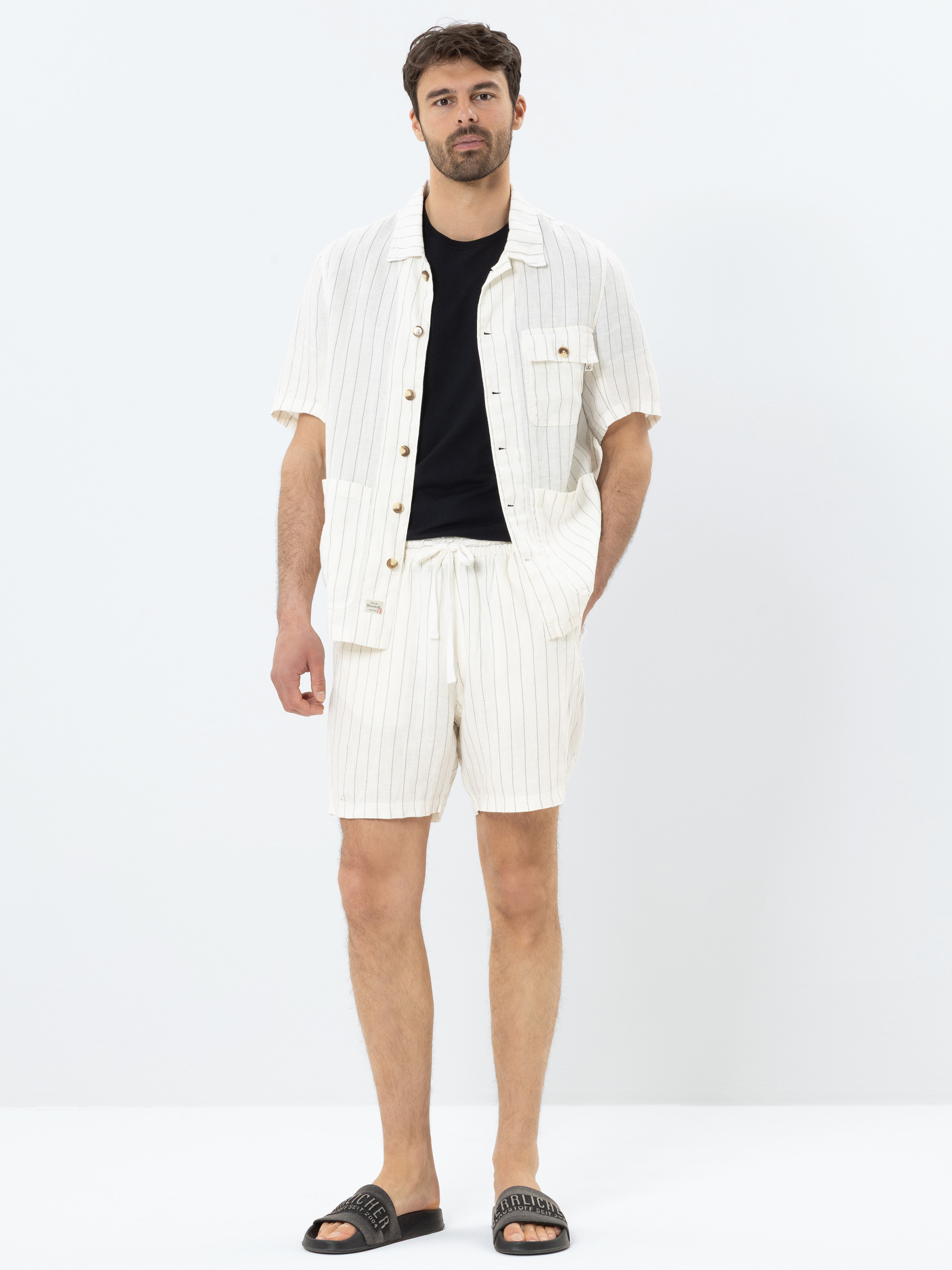 Ein Mann steht mit dem Gesicht nach vorne und trägt ein schwarzes T-Shirt, ein cremefarbenes, kurzärmeliges Hemd mit vertikalen Streifen, passende Shorts und schwarze Slide-Sandalen vor einem schlichten weißen Hintergrund.