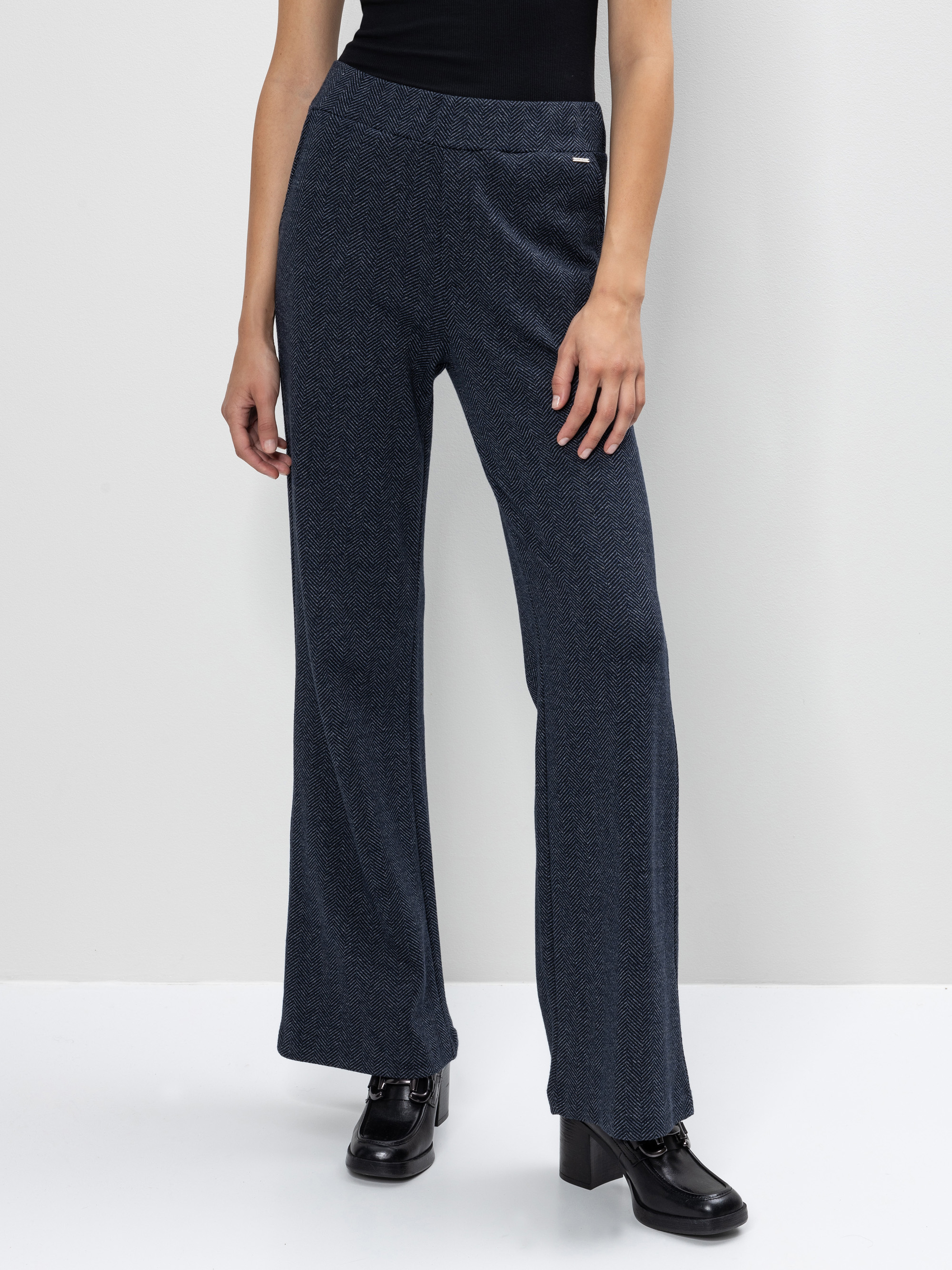 Eine Person, die eine dunkelblaue Hose mit weitem Bein, ein schwarzes ärmelloses Oberteil und schwarze hochhackige Schuhe trägt, steht vor einem hellgrauen Hintergrund. Nur der Körper von den Schultern abwärts ist sichtbar.