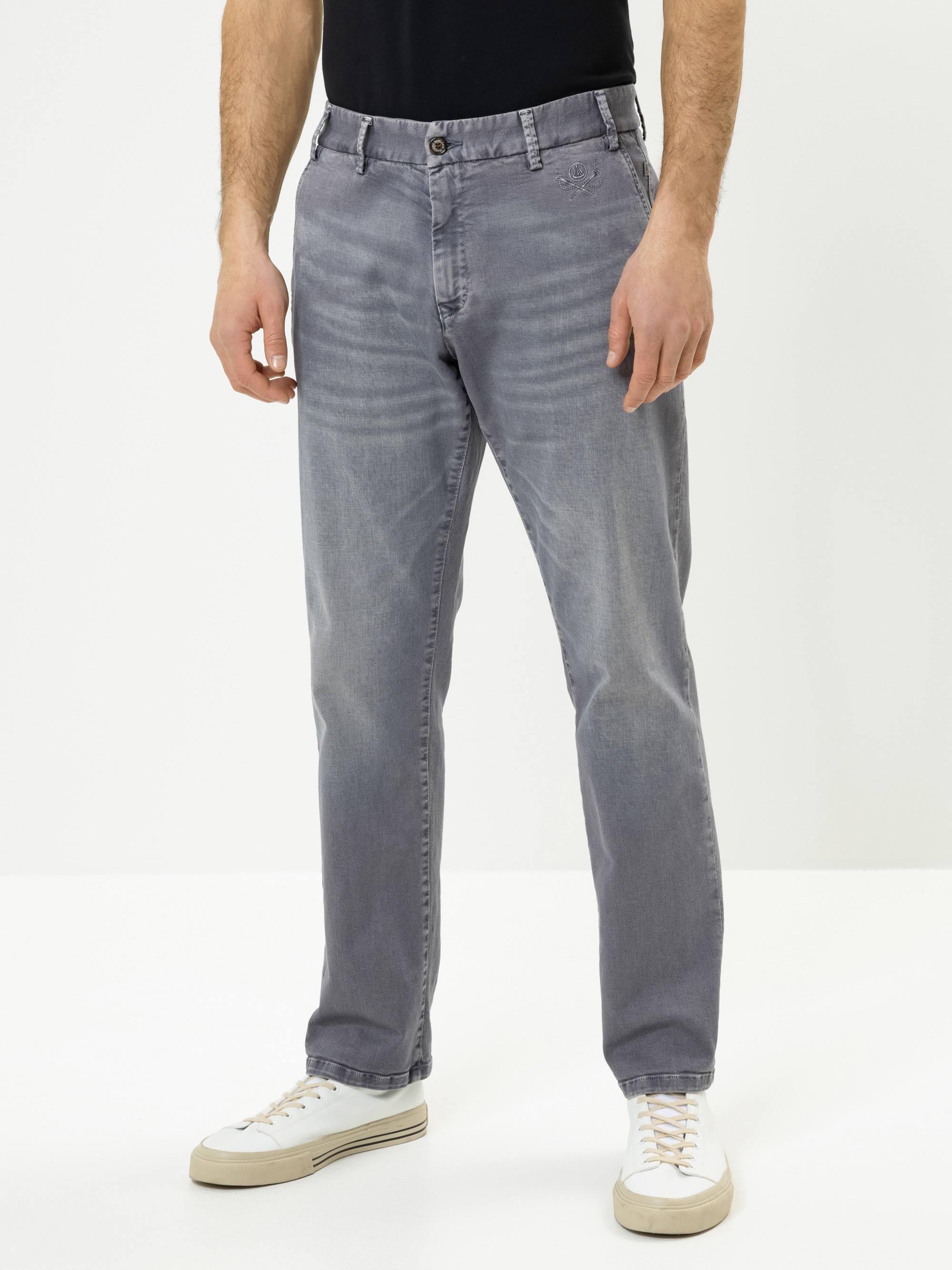 Eine Person, die ein schwarzes Hemd, verblasste graue Jeans und weiße Turnschuhe trägt, steht vor einem schlichten weißen Hintergrund. Das Bild zeigt die Person von den Schultern bis zu den Füßen.