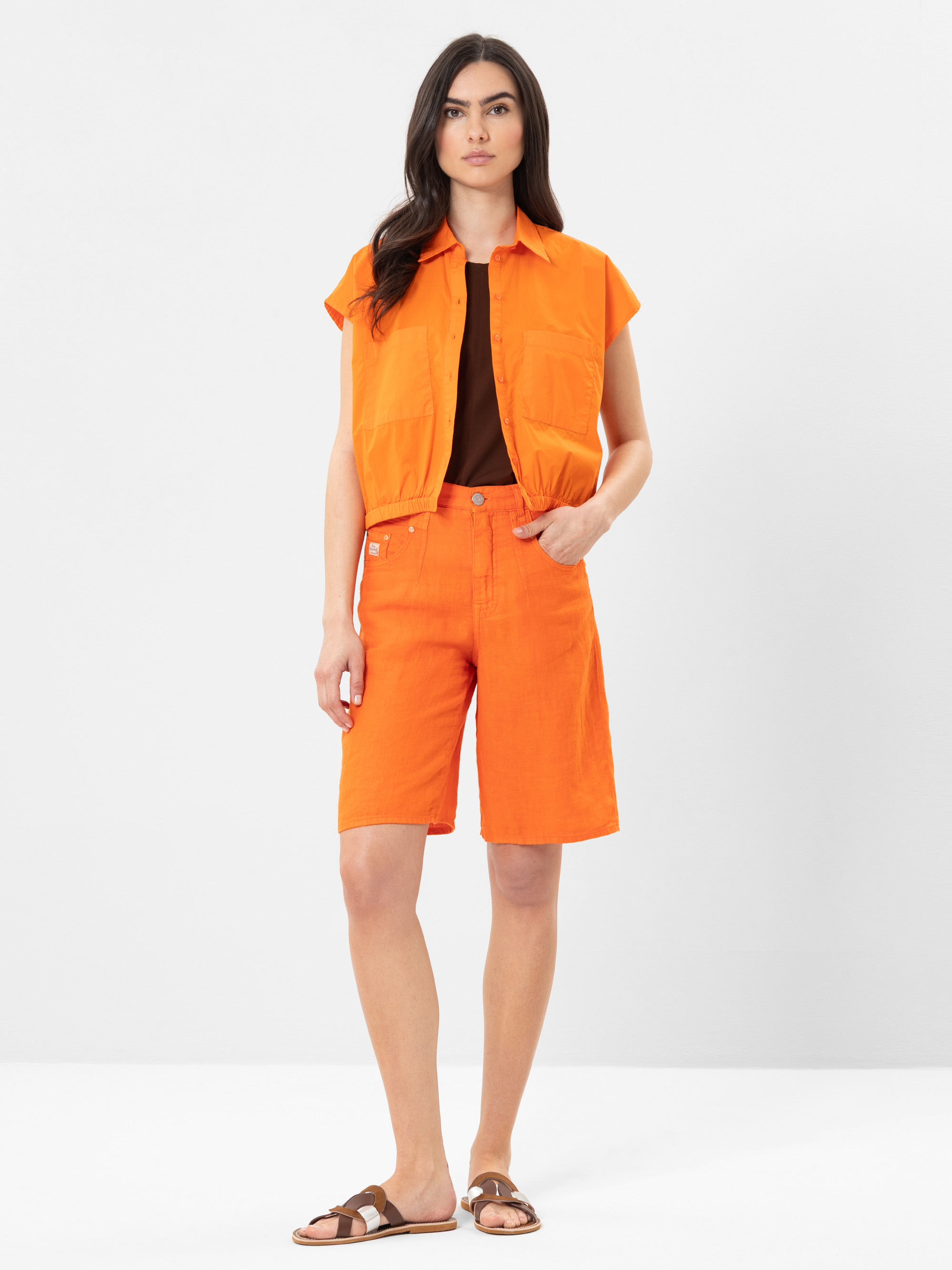 Eine Frau steht vor einem weißen Hintergrund und trägt ein orangefarbenes, ärmelloses Button-up-Shirt, dazu passende orangefarbene, knielange Shorts, ein dunkelbraunes Top und braune Sandalen.