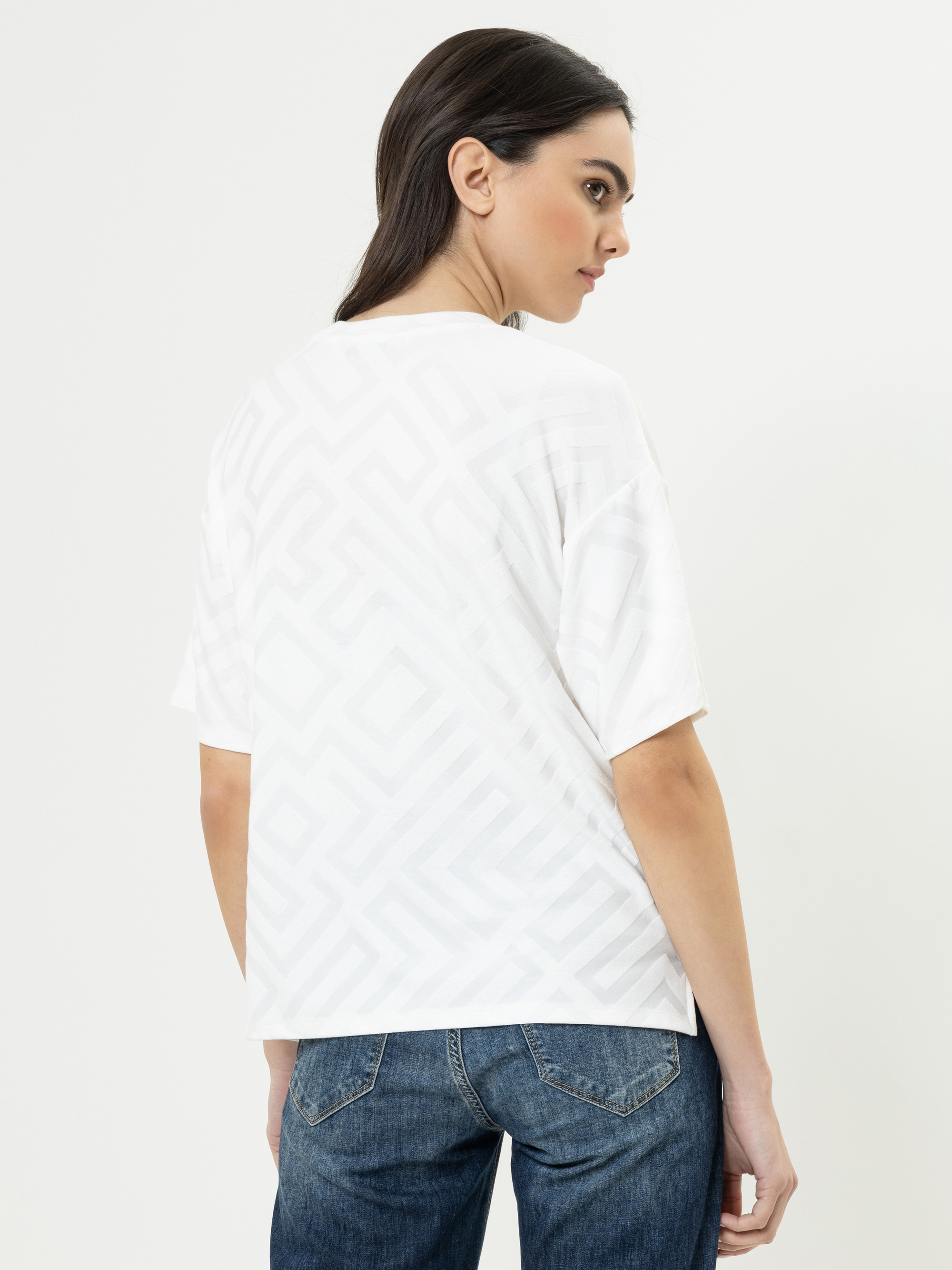 Eine Frau mit langen dunklen Haaren trägt ein weißes gemustertes T-Shirt und blaue Jeans. Sie steht mit dem Rücken zur Kamera und dreht ihren Kopf nach links vor einem schlichten weißen Hintergrund.