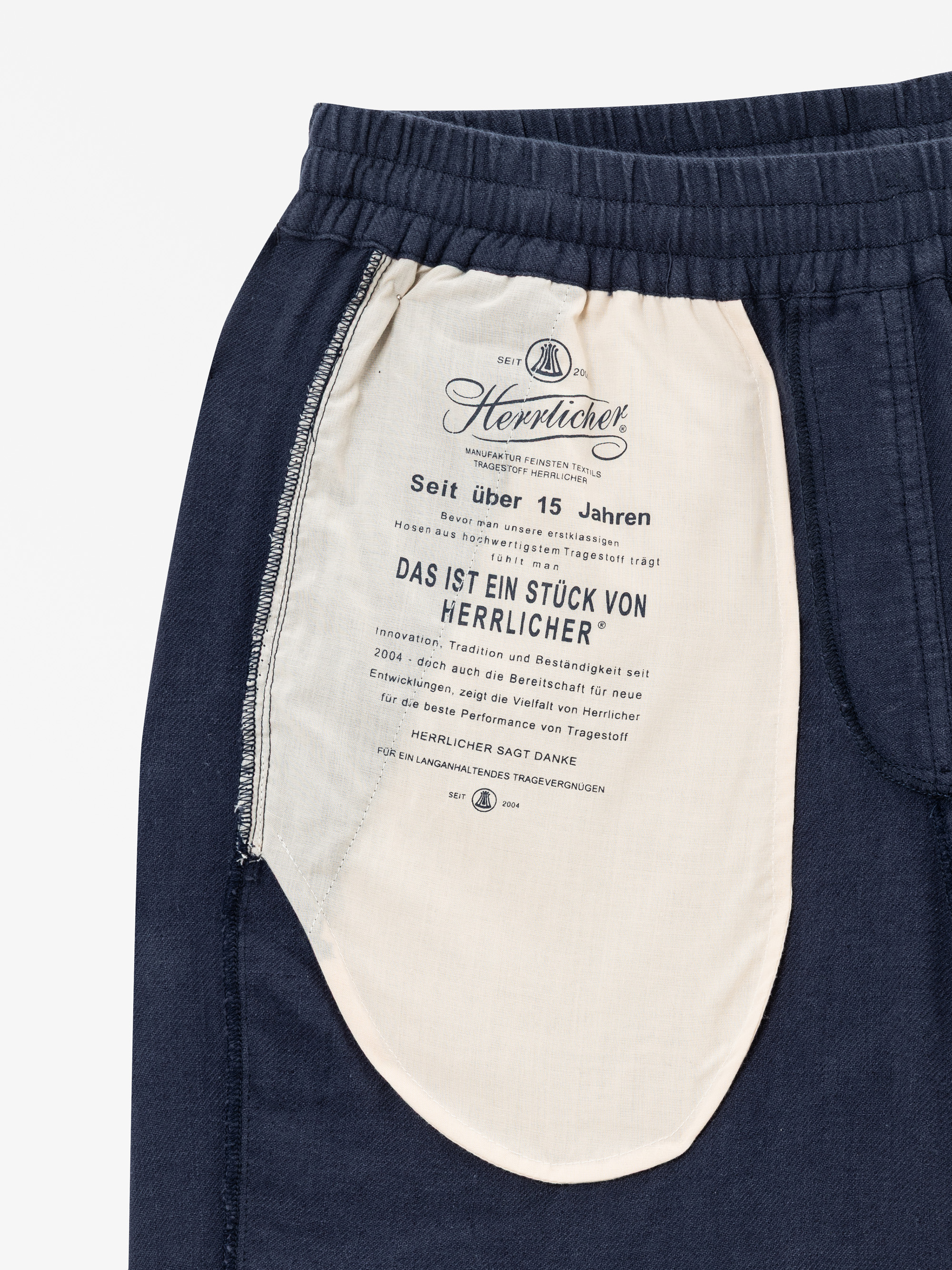 Nahaufnahme einer marineblauen Hose mit einem cremefarbenen Taschenfutter. Die Innenseite der Tasche ist mit schwarzem Text in deutscher Sprache bedruckt, der das Branding und die Pflegehinweise des Bekleidungsunternehmens Herrlicher enthält.