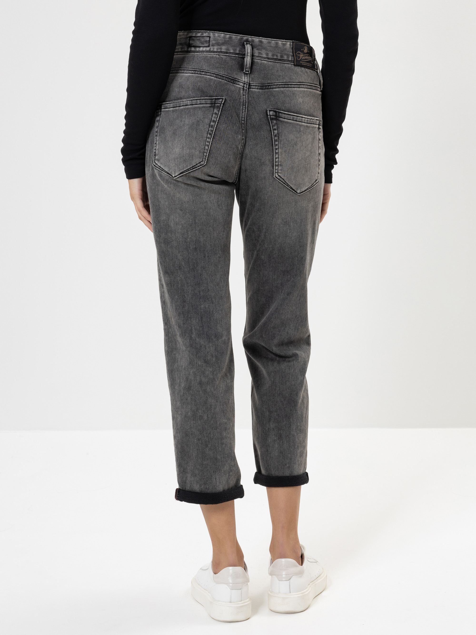 Eine Person steht mit dem Gesicht zur Wand und trägt eine schwarze, verwaschene Jeans mit sichtbaren Nähten und umgeschlagenen Bündchen, ein schwarzes, langärmeliges Oberteil und weiße Turnschuhe vor einem schlichten weißen Hintergrund.