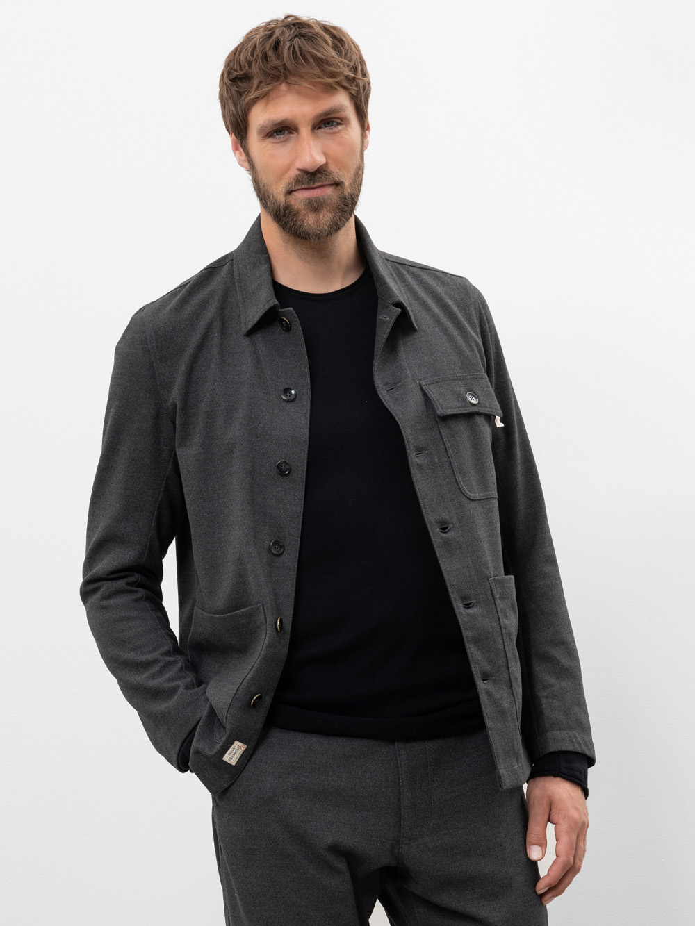 Ein Mann mit kurzen braunen Haaren und einem Bart trägt eine anthrazitfarbene Jacke über einem schwarzen Hemd und einer passenden grauen Hose. Er steht vor einem schlichten weißen Hintergrund, hat eine Hand in der Tasche und lächelt leicht.