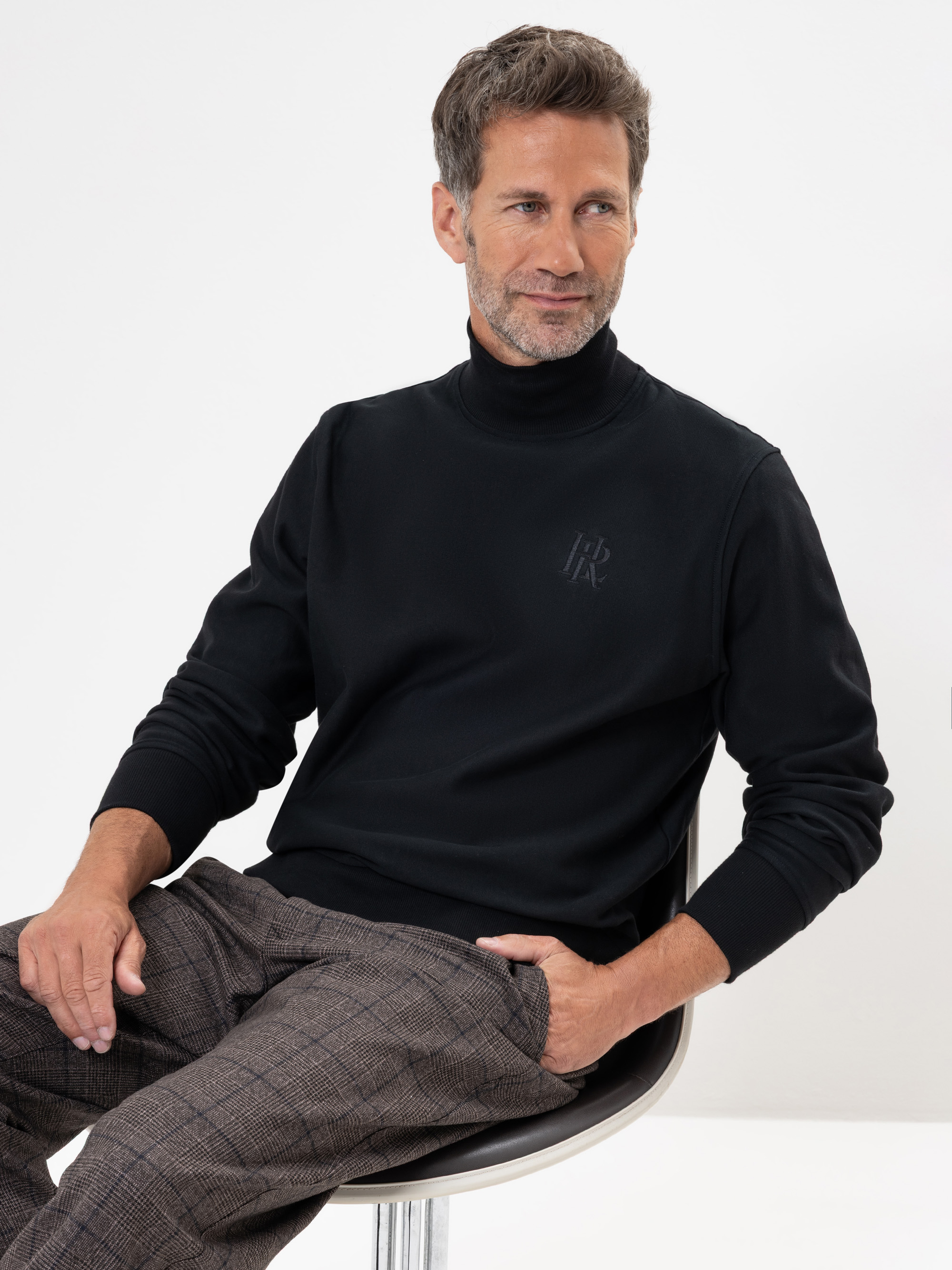 Ein Mann mit kurzen, braunen Haaren und einem Bart sitzt auf einem Stuhl, trägt einen schwarzen Rollkragenpullover und eine grau karierte Hose und blickt leicht zur Seite mit einem entspannten Ausdruck vor einem schlichten weißen Hintergrund.