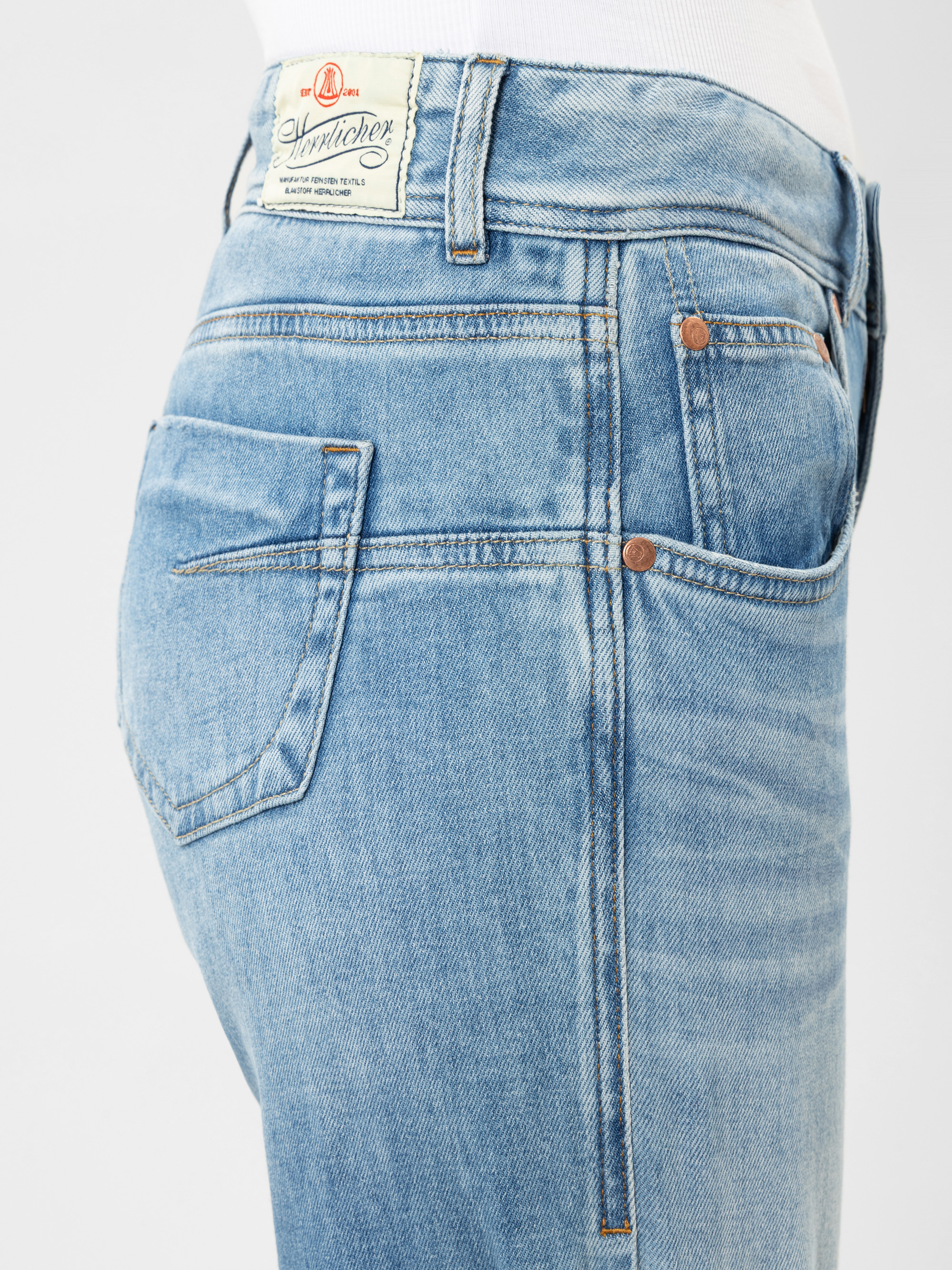 Nahaufnahme einer Person, die eine hellblaue Jeans mit einem sichtbaren weißen Etikett am Bund und kupferfarbenen Nieten an den Taschen trägt. Die Person trägt ein weißes Hemd, das in die Jeans gesteckt ist.