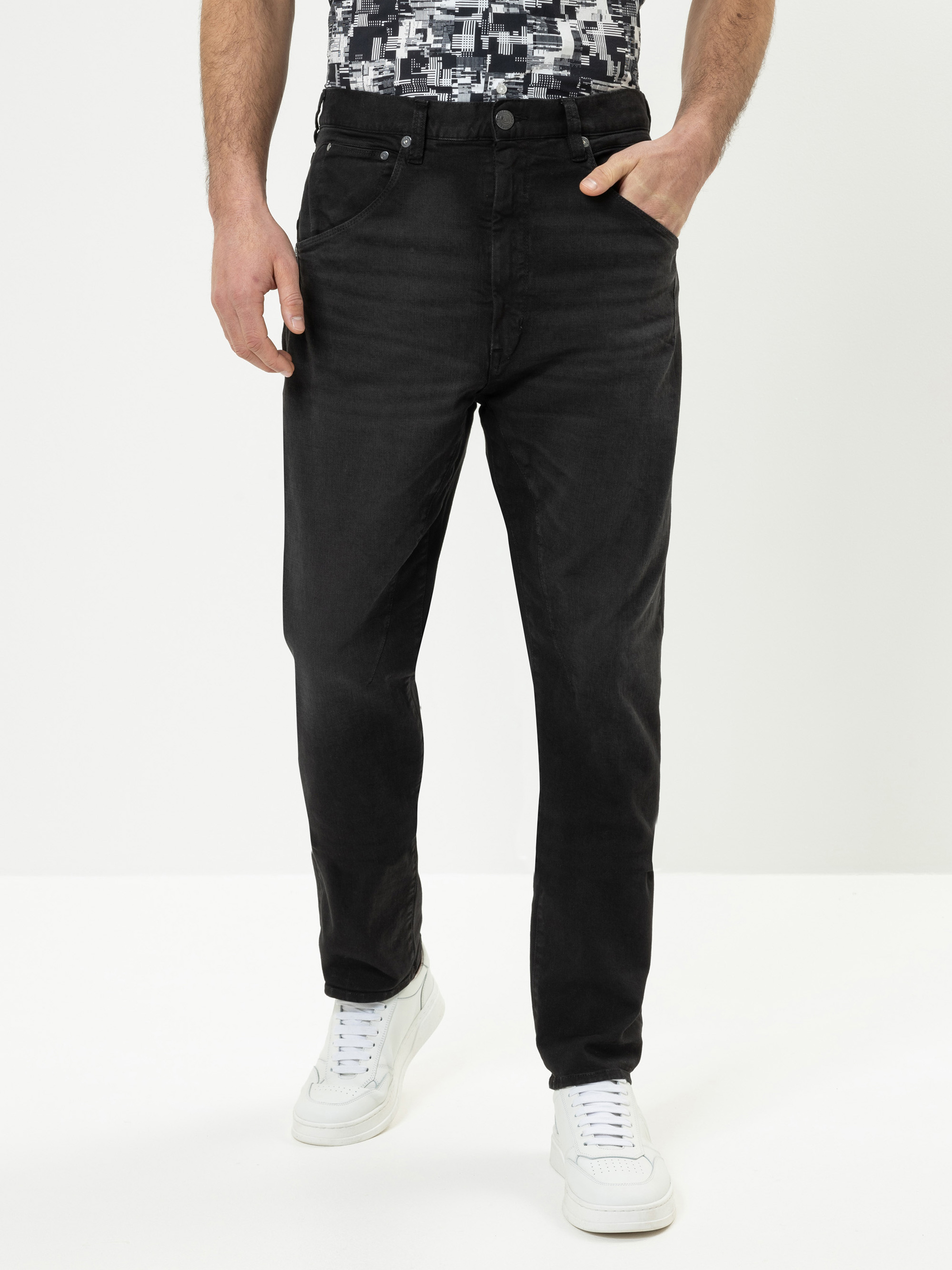 Eine Person, die schwarze Jeans, ein gemustertes Hemd und weiße Turnschuhe trägt, steht vor einem schlichten weißen Hintergrund und hat eine Hand in einer Tasche. Das Bild zeigt die Person von den Schultern bis zu den Füßen.