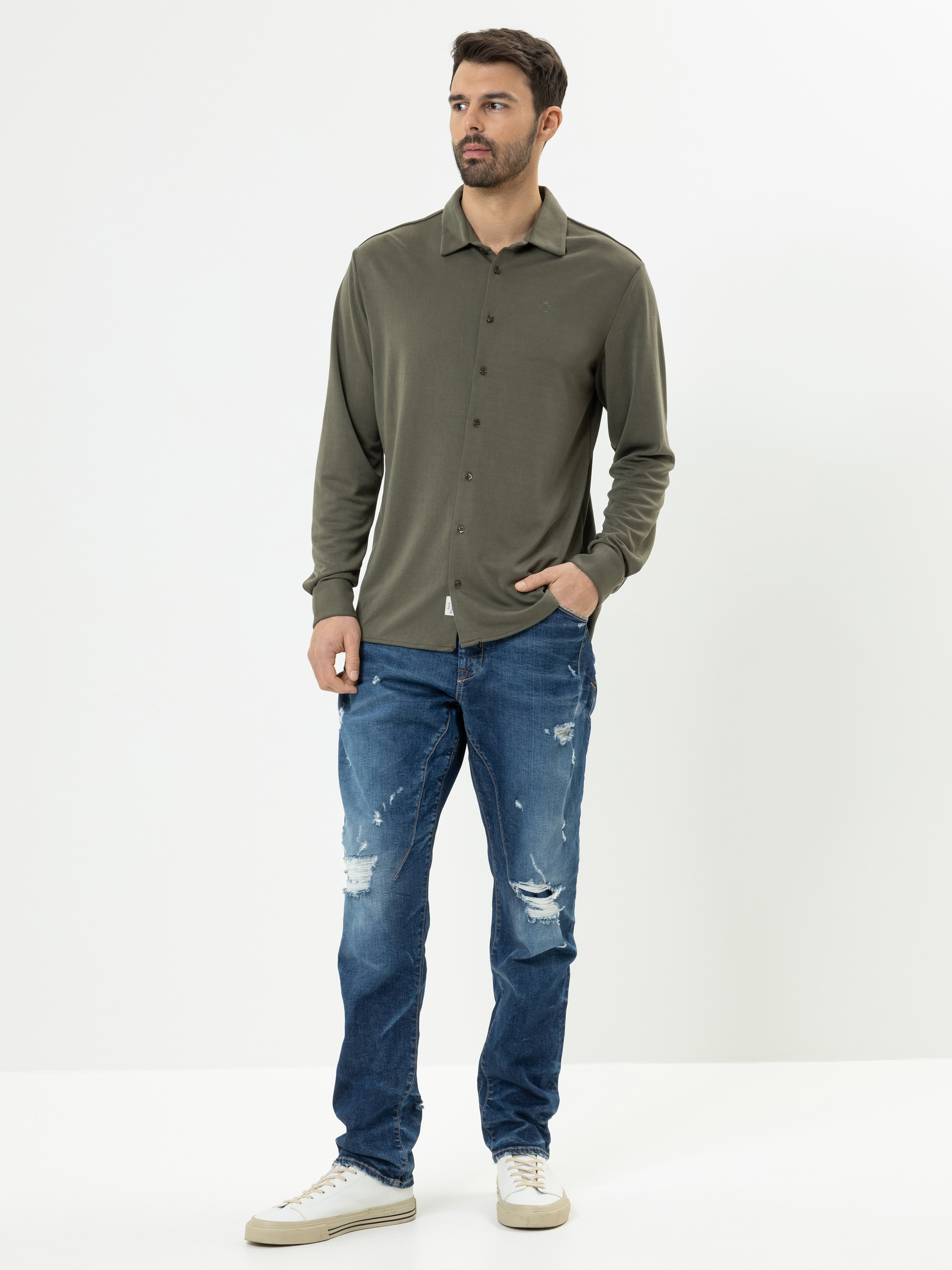 Ein Mann steht vor einem schlichten weißen Hintergrund und trägt ein grünes Button-up-Hemd, abgewetzte Bluejeans und weiße Turnschuhe. Er hat kurzes dunkles Haar, einen Bart und schaut leicht nach links, die Hände in den Taschen.