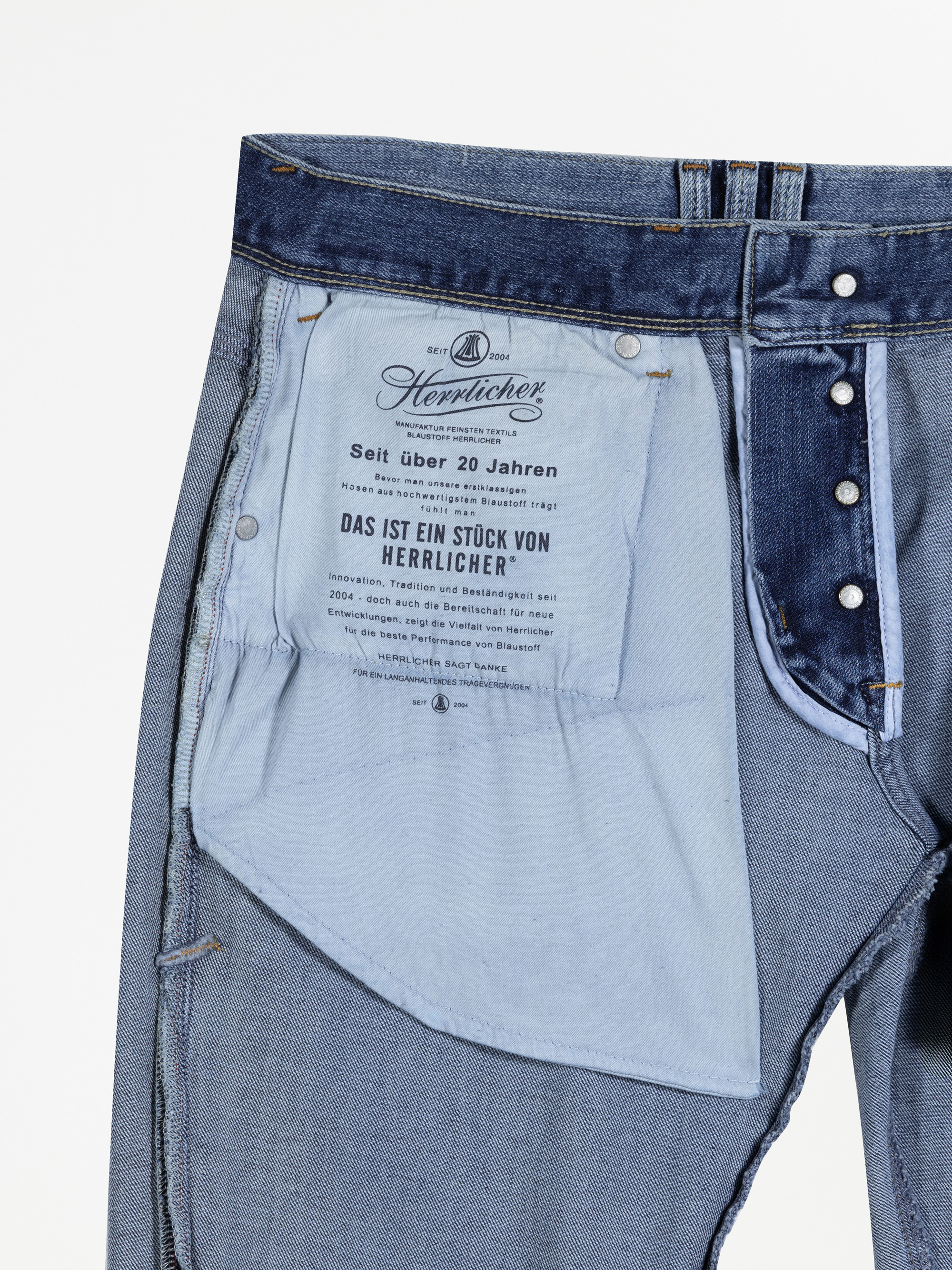 Innenansicht einer blauen Jeans, die den weißen Stoff der Innentasche mit deutschem Text, silbernen Nieten und sichtbaren Nähten am Bund und Saum zeigt.