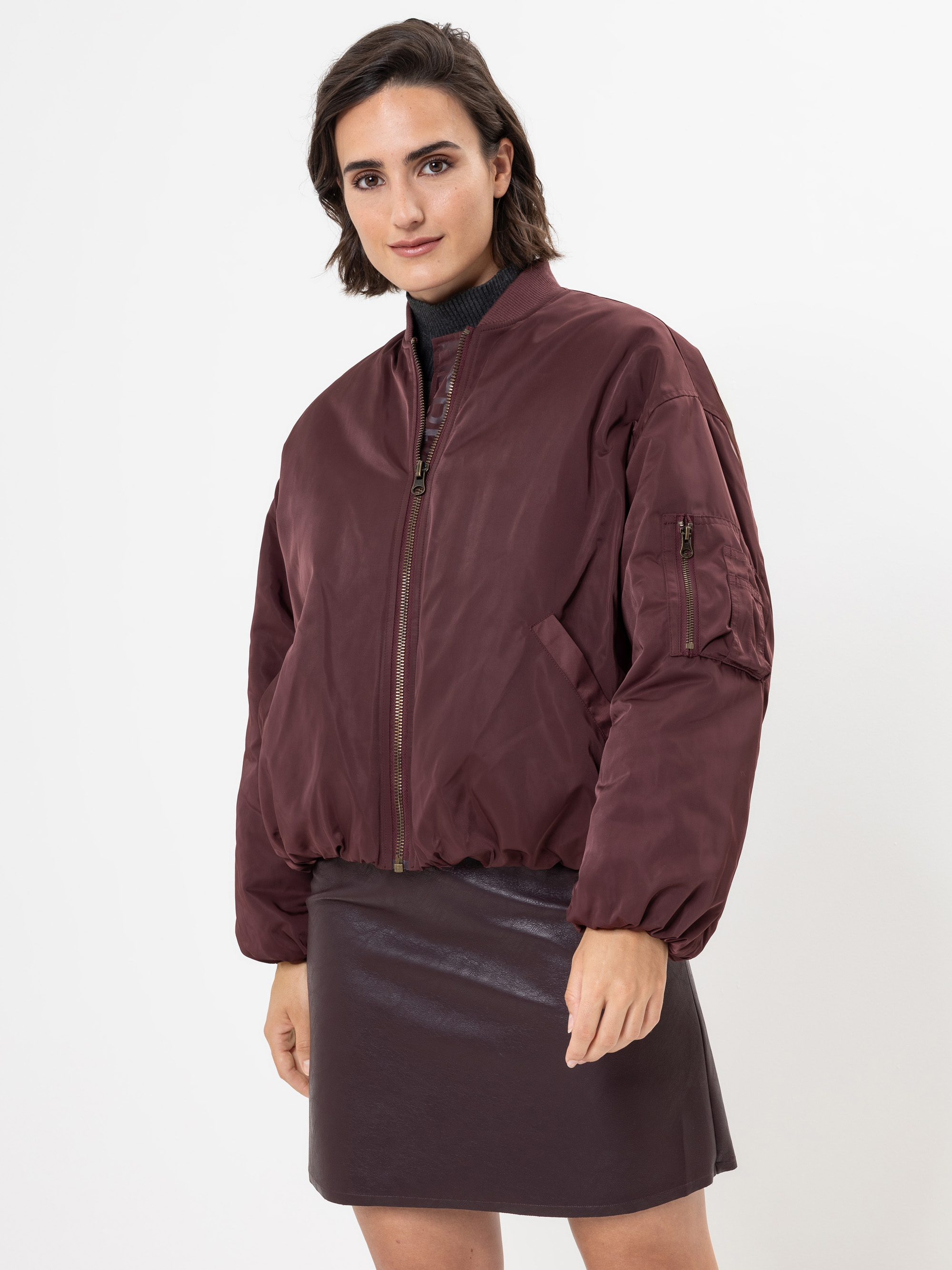 Eine Frau, die eine kastanienbraune Bomberjacke über einem dunkelbraunen Lederrock trägt, steht vor einem schlichten weißen Hintergrund und schaut selbstbewusst in die Kamera.