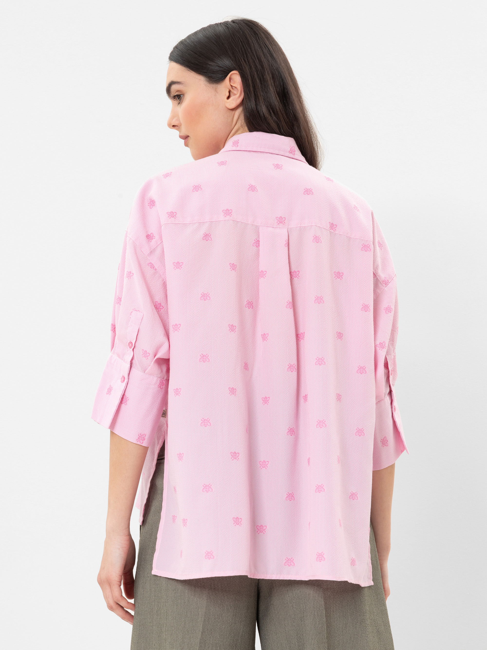 Eine Frau mit langen braunen Haaren trägt ein locker sitzendes, hellrosafarbenes Button-up-Hemd mit hochgekrempelten Ärmeln und einem dezenten Muster, dazu eine olivgrüne Hose. Sie blickt von der Kamera weg und steht vor einem schlichten weißen Hinter