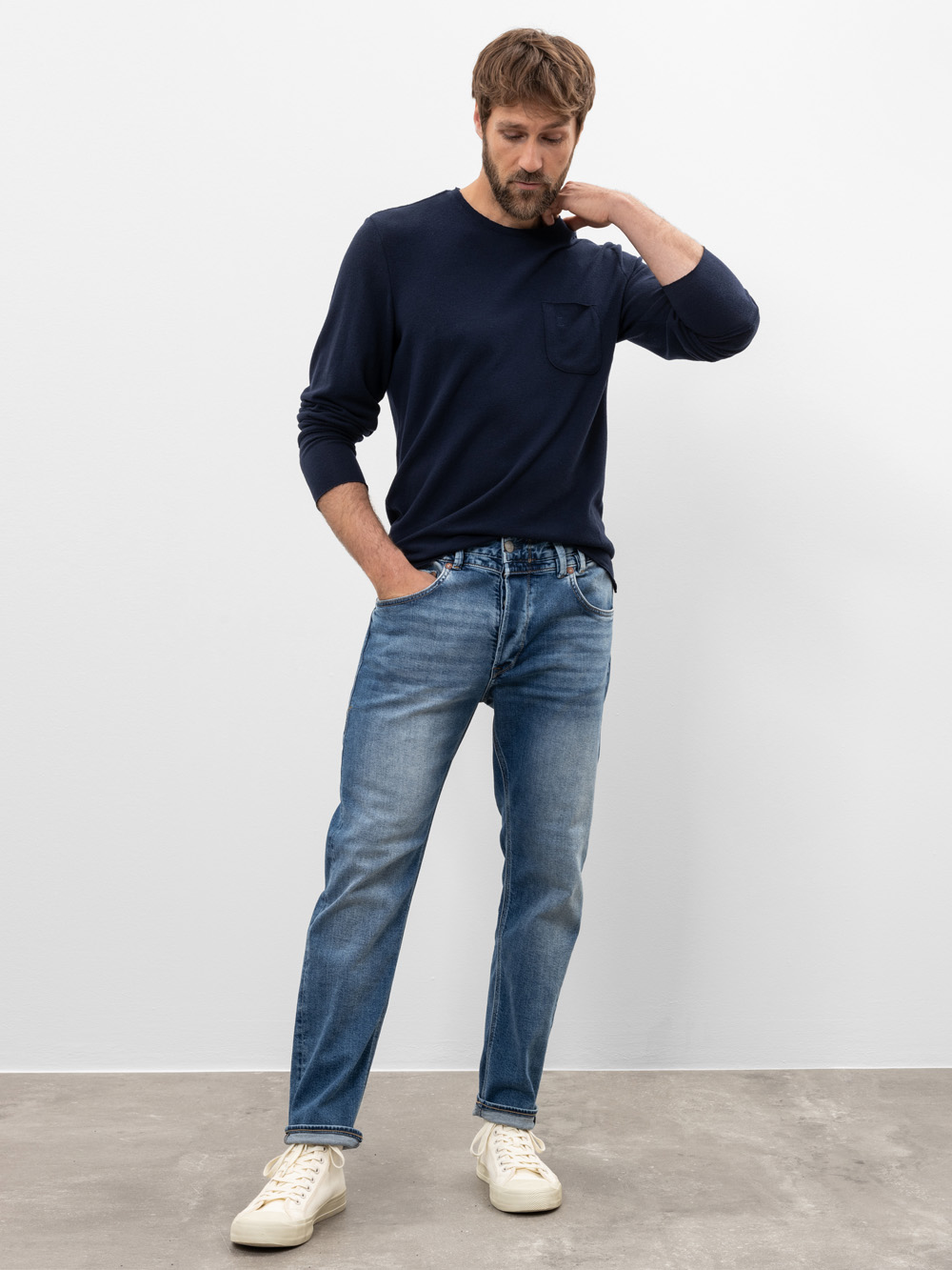 Ein Mann steht in einem Innenraum vor einer schlichten weißen Wand. Er trägt ein marineblaues langärmeliges Hemd, blaue Jeans und weiße Turnschuhe. Er hat eine Hand in der Hosentasche, die andere berührt seinen Nacken und schaut nach unten.